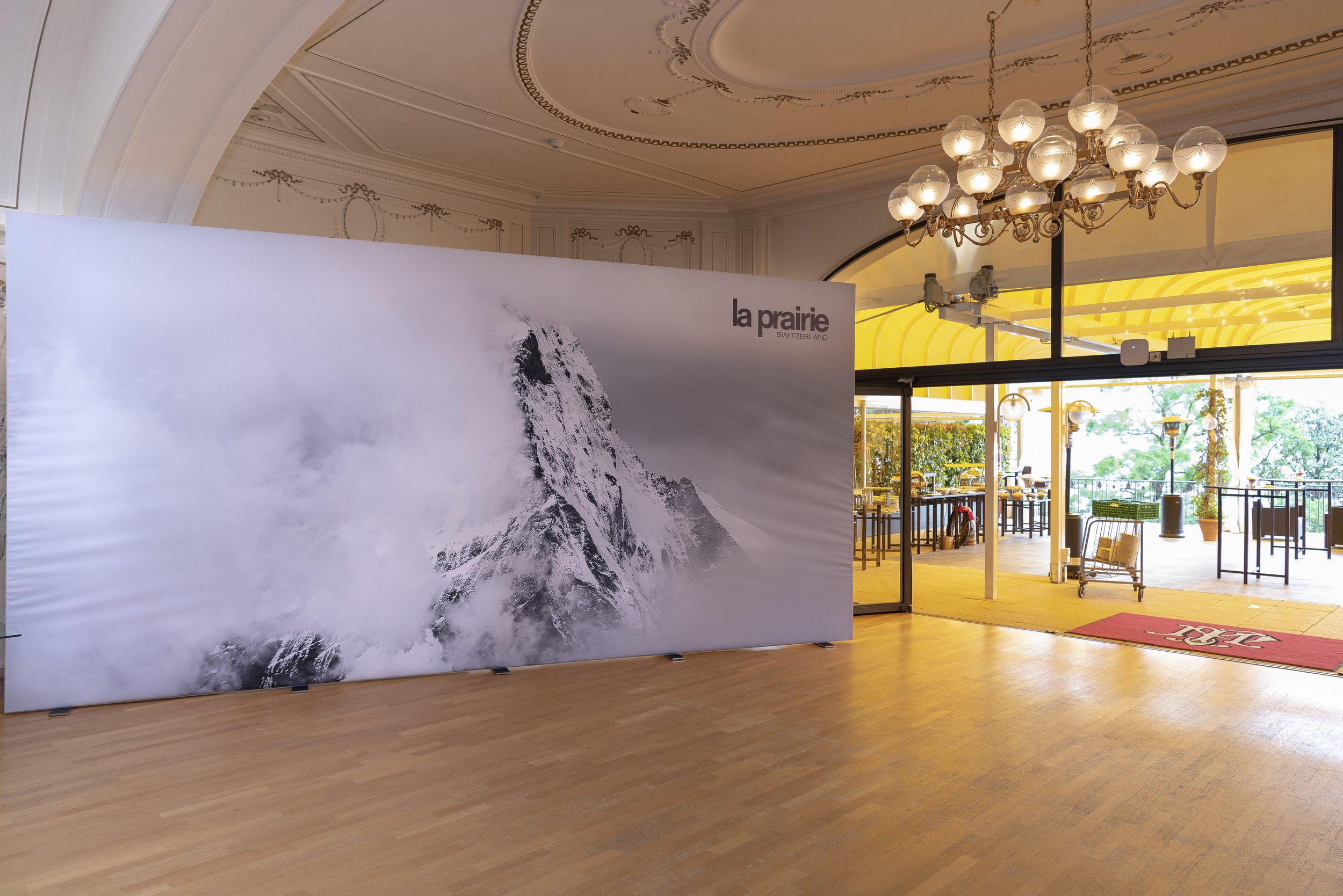 Une grande affiche représentant la montagne Matterhorn dans les Alpes suisses, située à l'intérieur d'une salle avec un plancher en bois, une chandelier suspendu au plafond et une porte qui mène à un espace extérieur lumineux avec des tables et des c