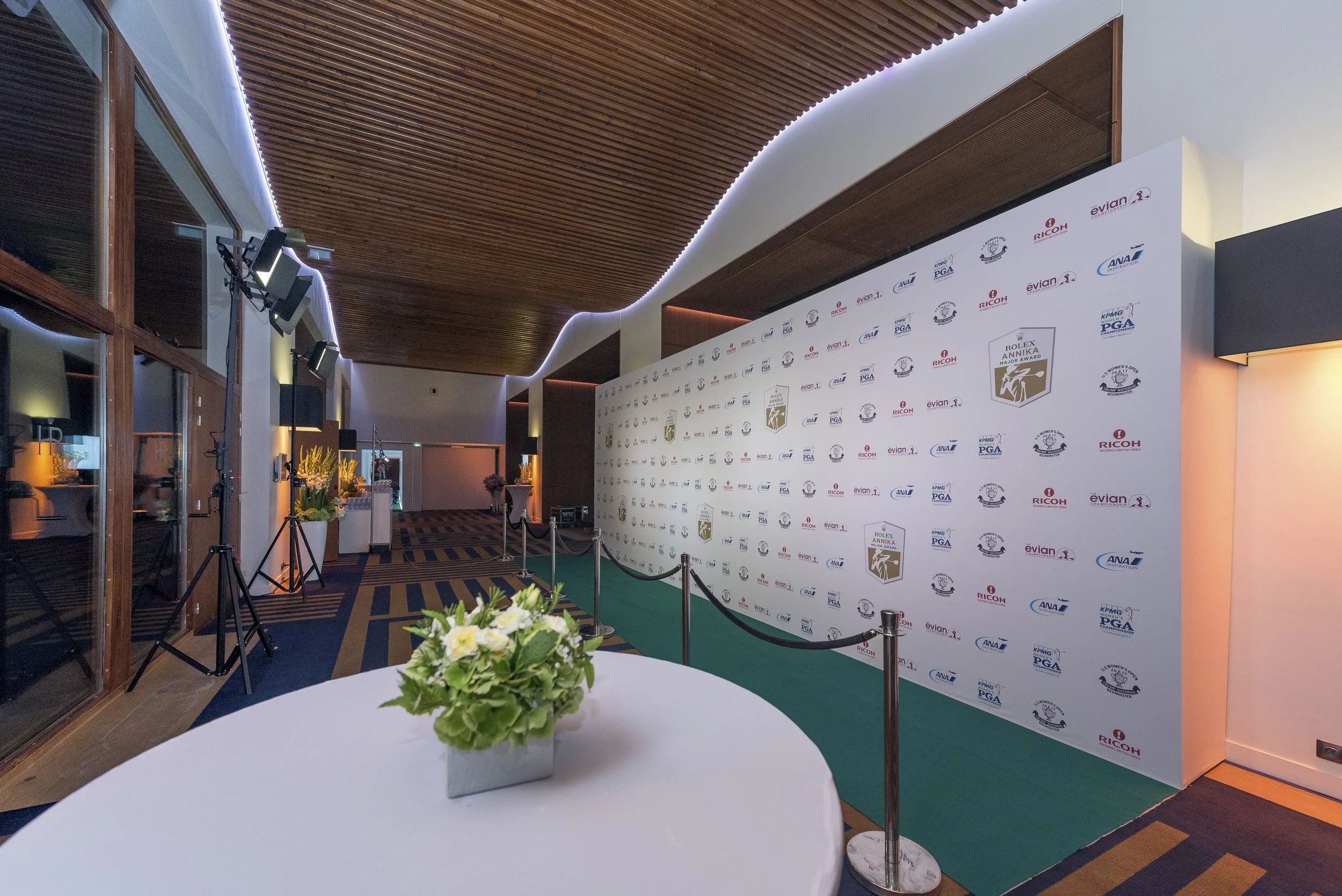 Espace de conférence ou événement avec un tapis vert, un fond avec logos et une table avec un bouquet de fleurs au premier plan