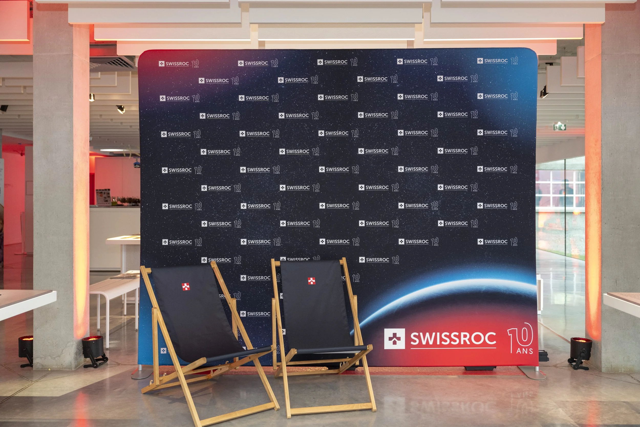 Deux chaises longues en bois avec bâches en tissu noir, placées devant une bannière promotionnelle de Swissroc, avec un fond représentant la terre vue de l'espace, célébrant les 10 ans de l'entreprise.