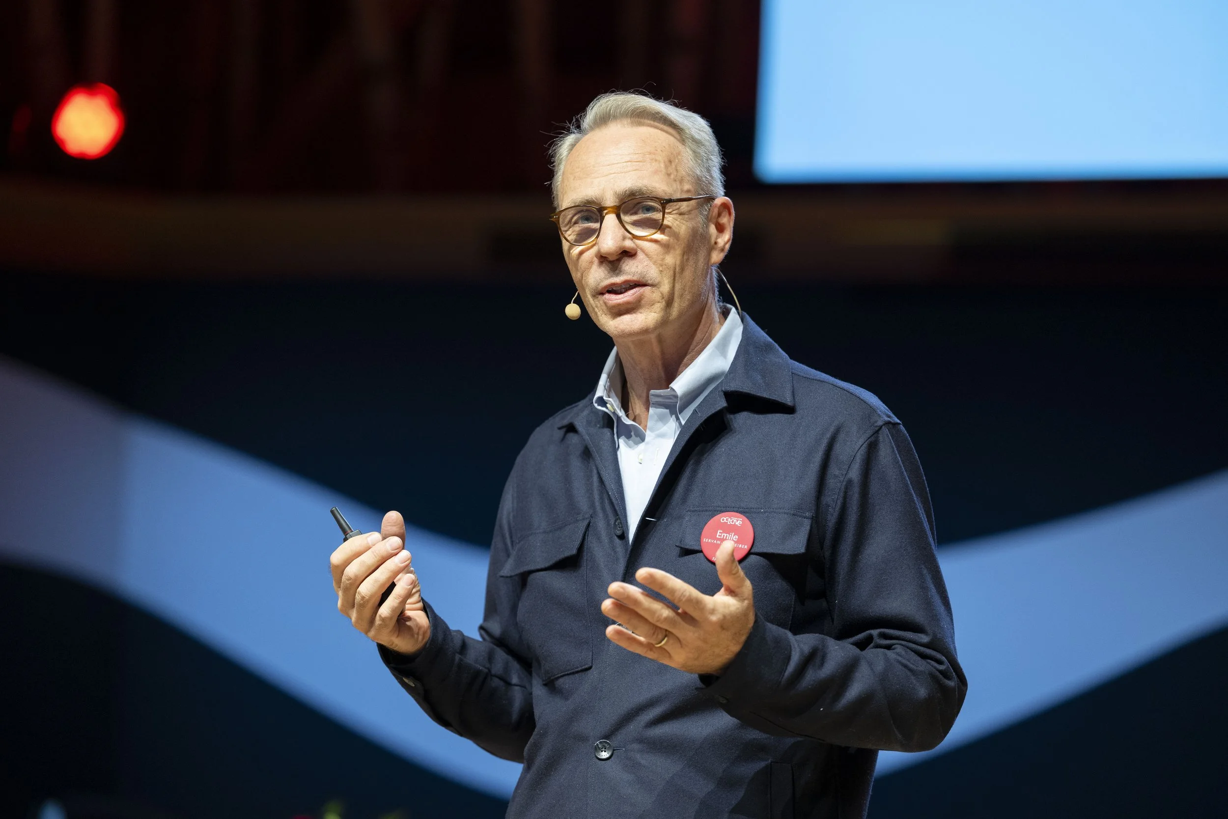 Un homme porte des lunettes, une chemise blanche et un veston foncé, parlant en public avec un micro intégré, tenant un pointeur dans la main. Il porte un badge rouge avec son nom, Emile. Le fond est sombre avec des formes bleues et un grand écran bl