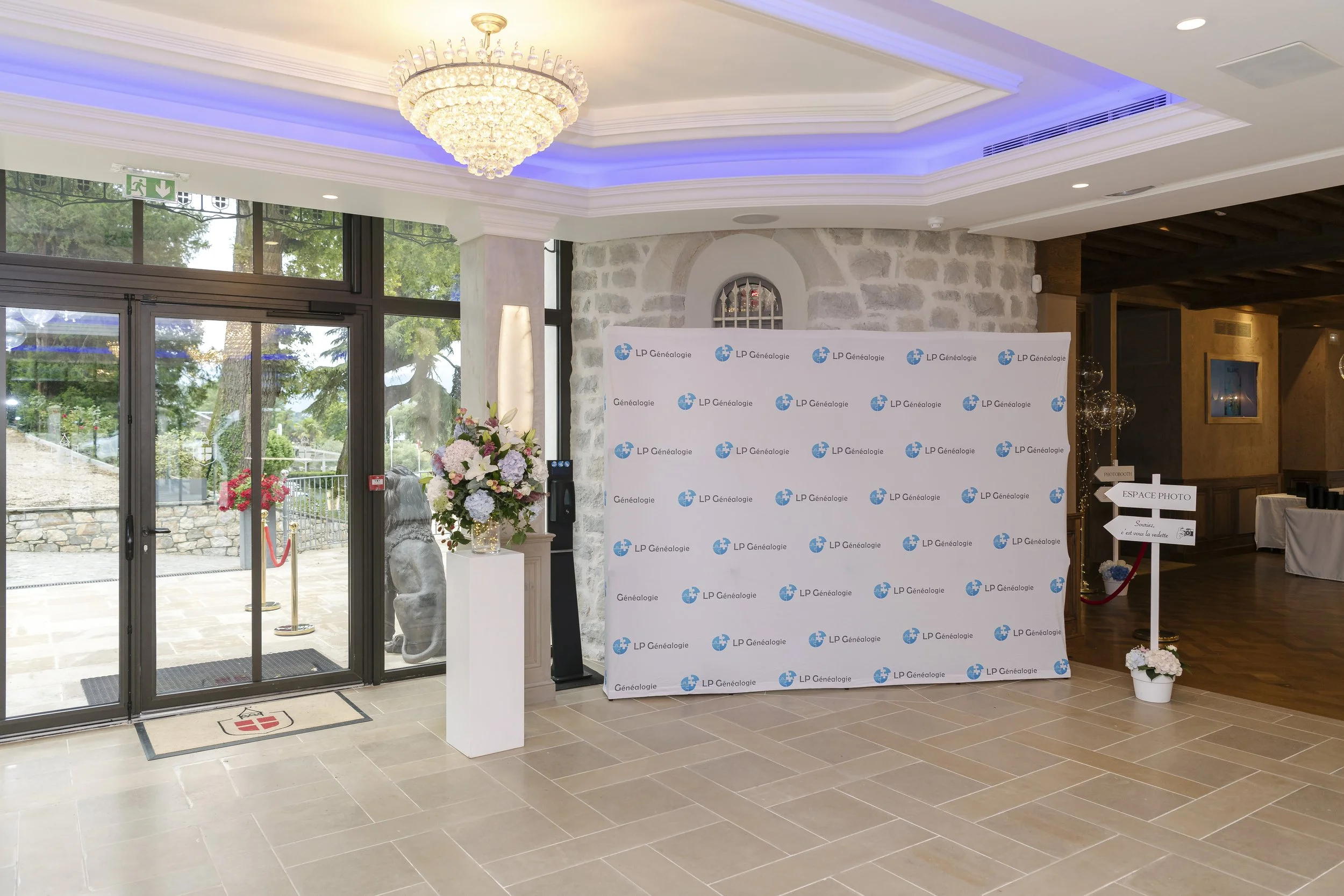 Entrée d'un événement avec une porte en verre, une grande affiche avec le logo 'LP Généalogie', un pique-fleur avec des fleurs, une statue de lion, un panneau directionnel 'Espace Photo', et un intérieur élégant avec un plafond lumineux et une décora
