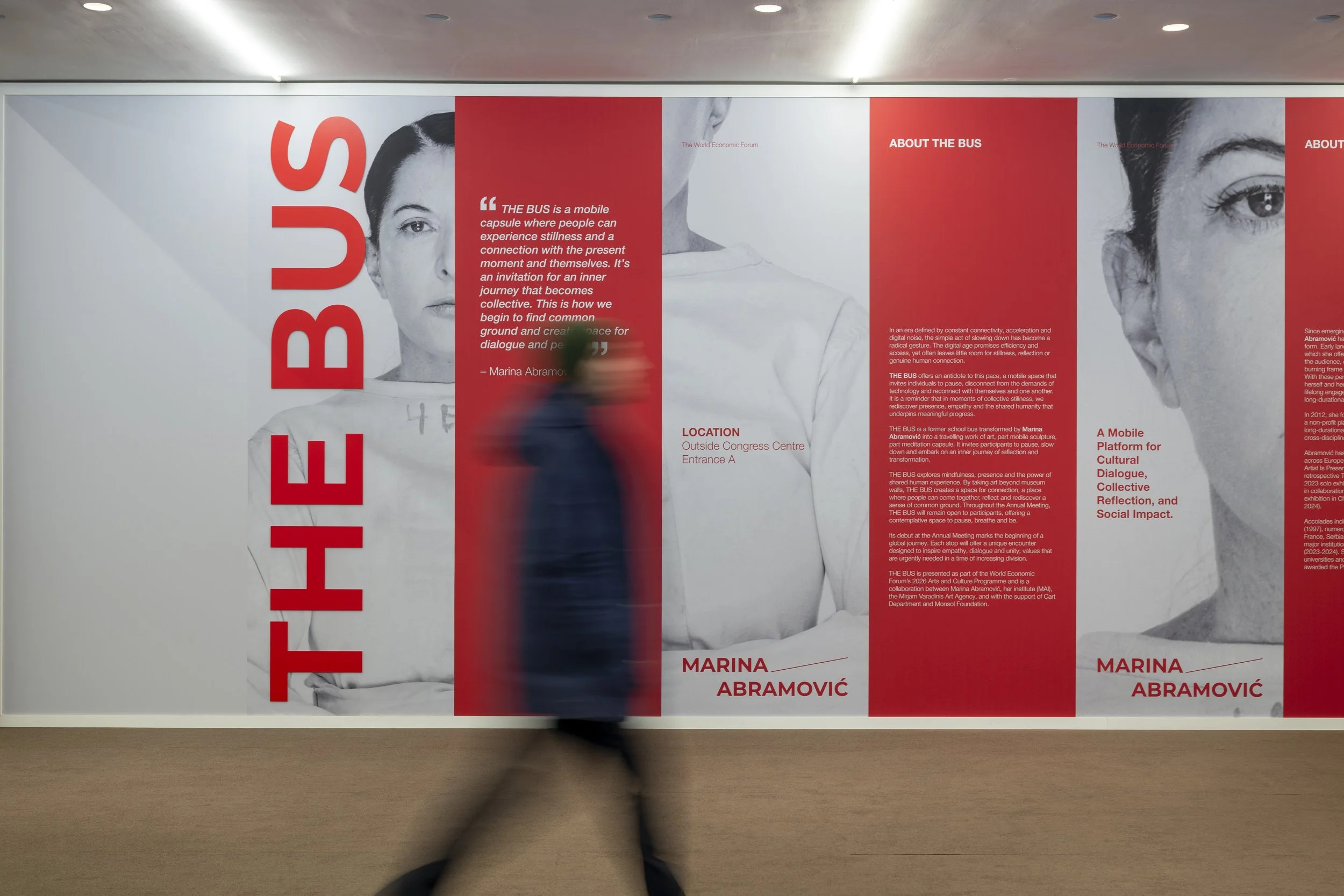 Une personne passant devant une grande affiche noir et blanc avec du texte rouge et blanc concernant Marina Abramović et un projet appelé 'The Bus'.