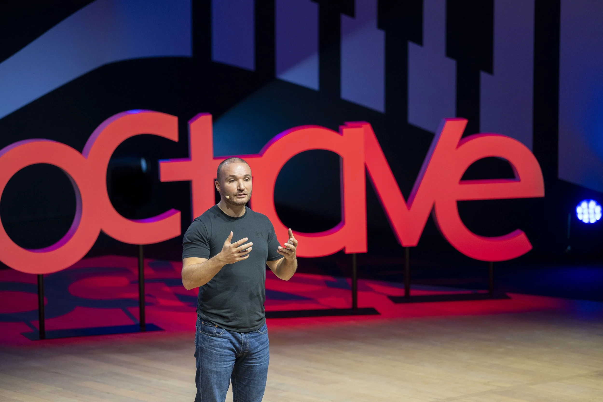 Un homme parle sur scène devant un grand panneau rouge avec le mot 'acte' inscrit dessus, lors d'une conférence TED.