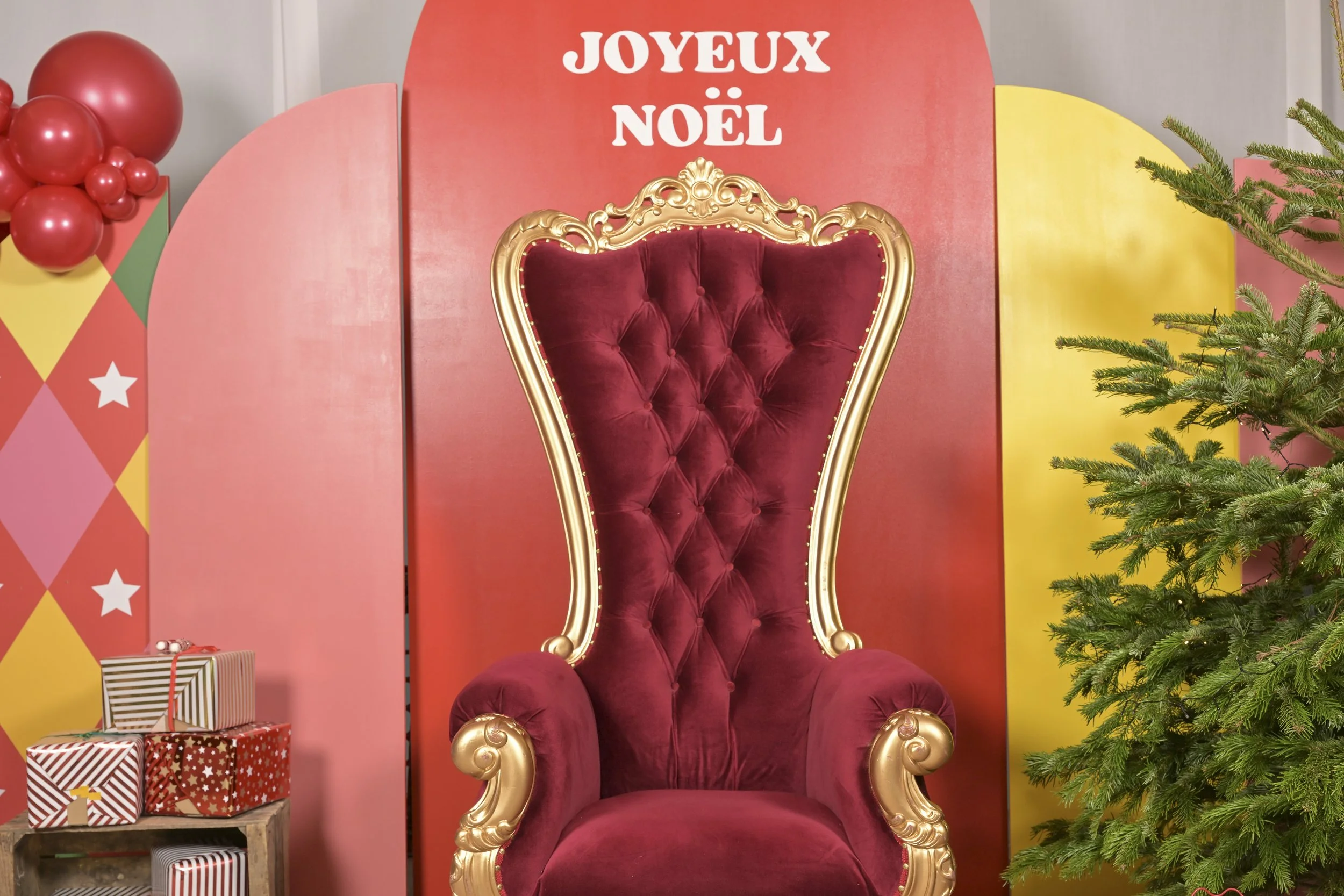 Chaise de roi royale en velours rouge avec cadre doré, décorée pour Noël, avec un sapin vert à droite et des cadeaux emballés en bas à gauche, en arrière-plan un panneau rouge avec 'Joyeux Noël'