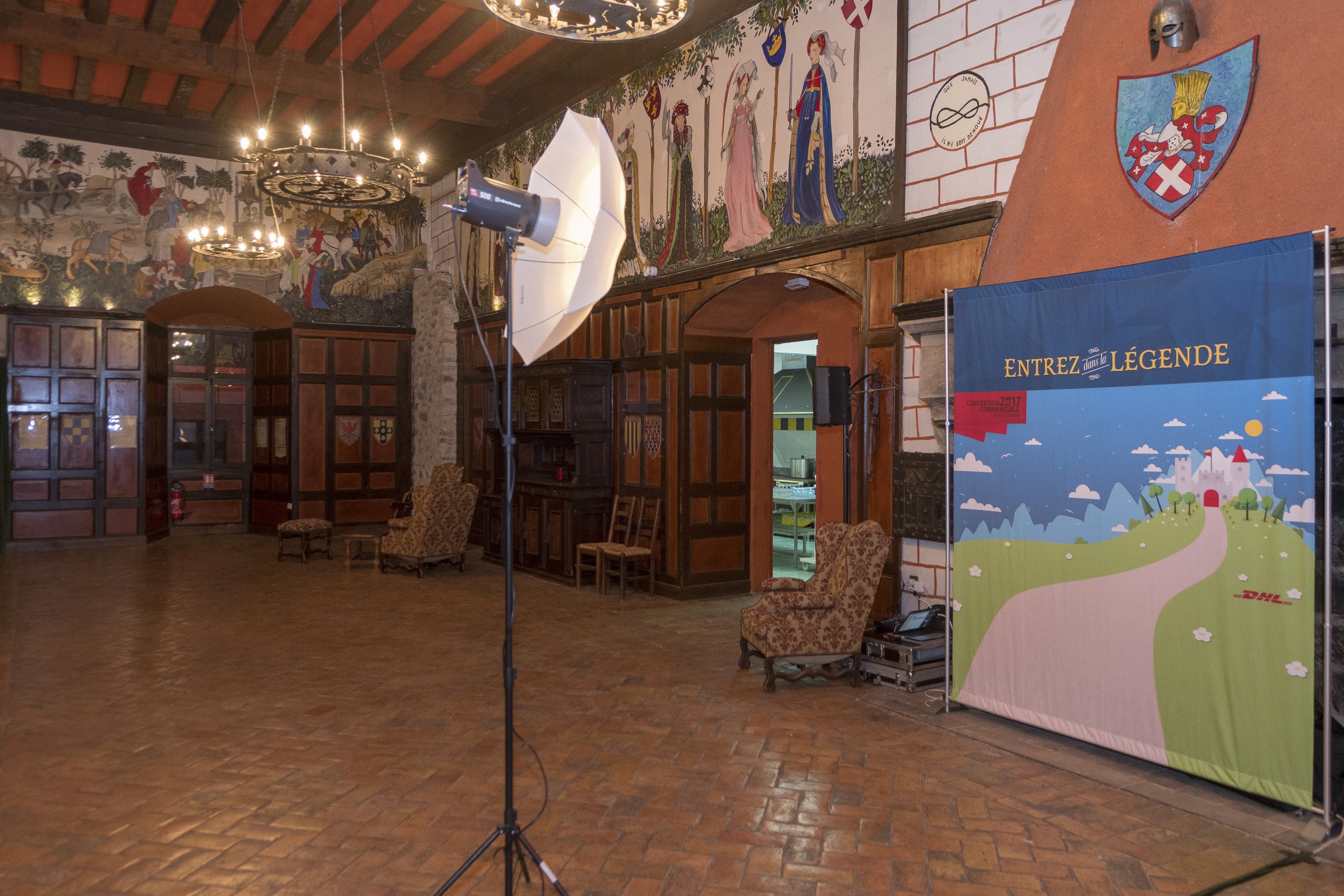 Salle de réception décorée avec des peintures médiévales, des armoiries, des fauteuils anciens, un panneau publicitaire avec la phrase "Entrez dans la légende" et un éclairage professionnel