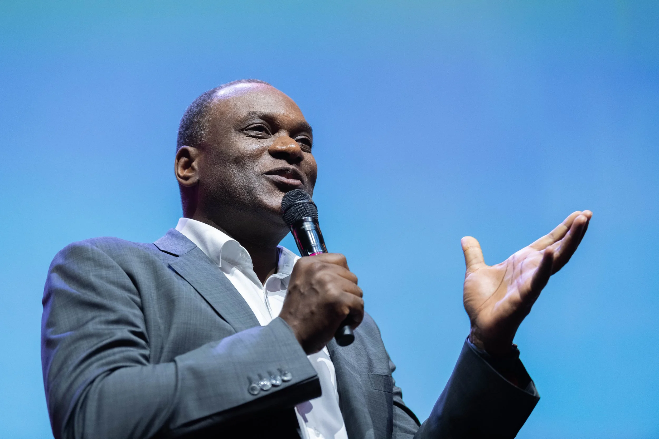 Homme en costume gris parlant en tenant un micro avec un fond bleu.