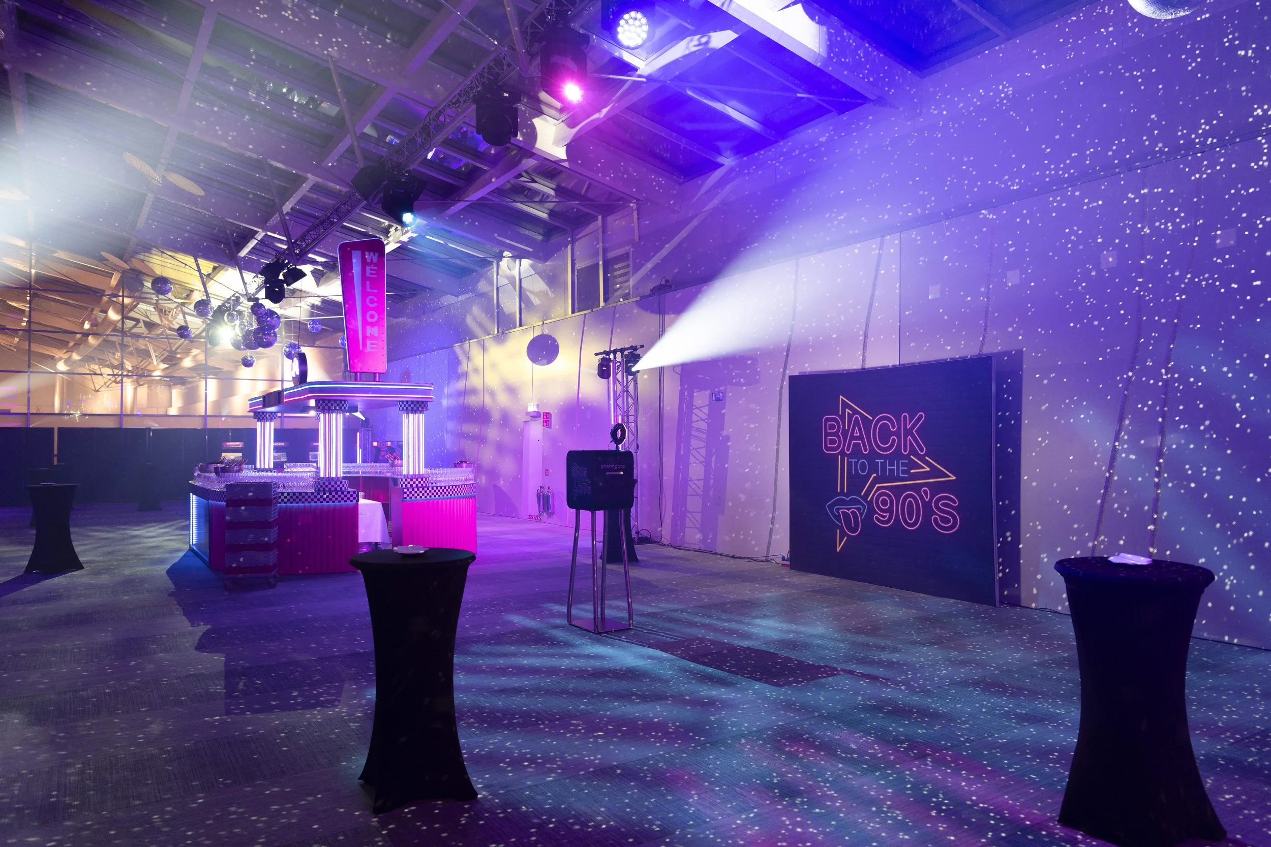 Une salle décorée pour une fête sur le thème des années 90 avec lumières colorées, une scène, une grande affiche neon indiquant 'Back to the 90s' et des tables hautes recouvertes de nappes noires.