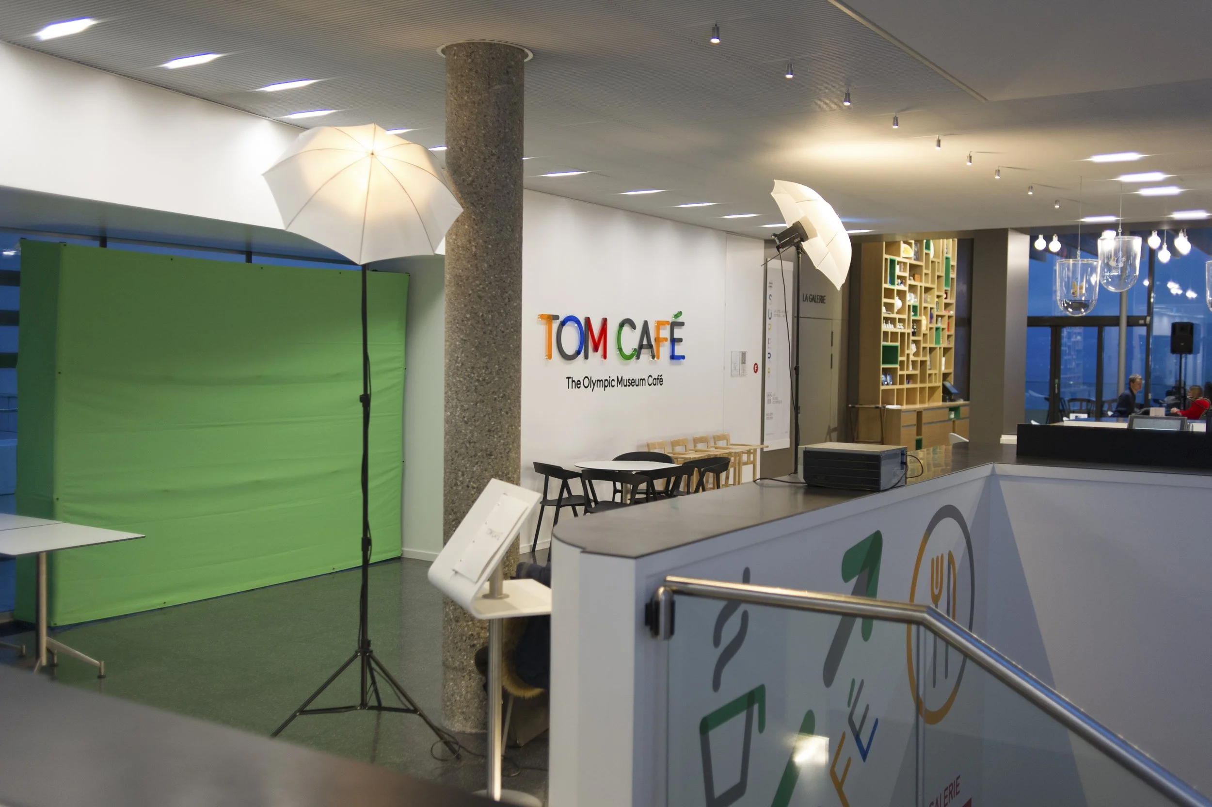 Intérieur du café du musée olympique, avec un arrière-plan vert pour photobooth, des parapluies blancs éclairés, une table avec des chaises noires, une libré de téléviseurs encadrés en bois, et des décorations modernes avec de grandes fenêtres en arr