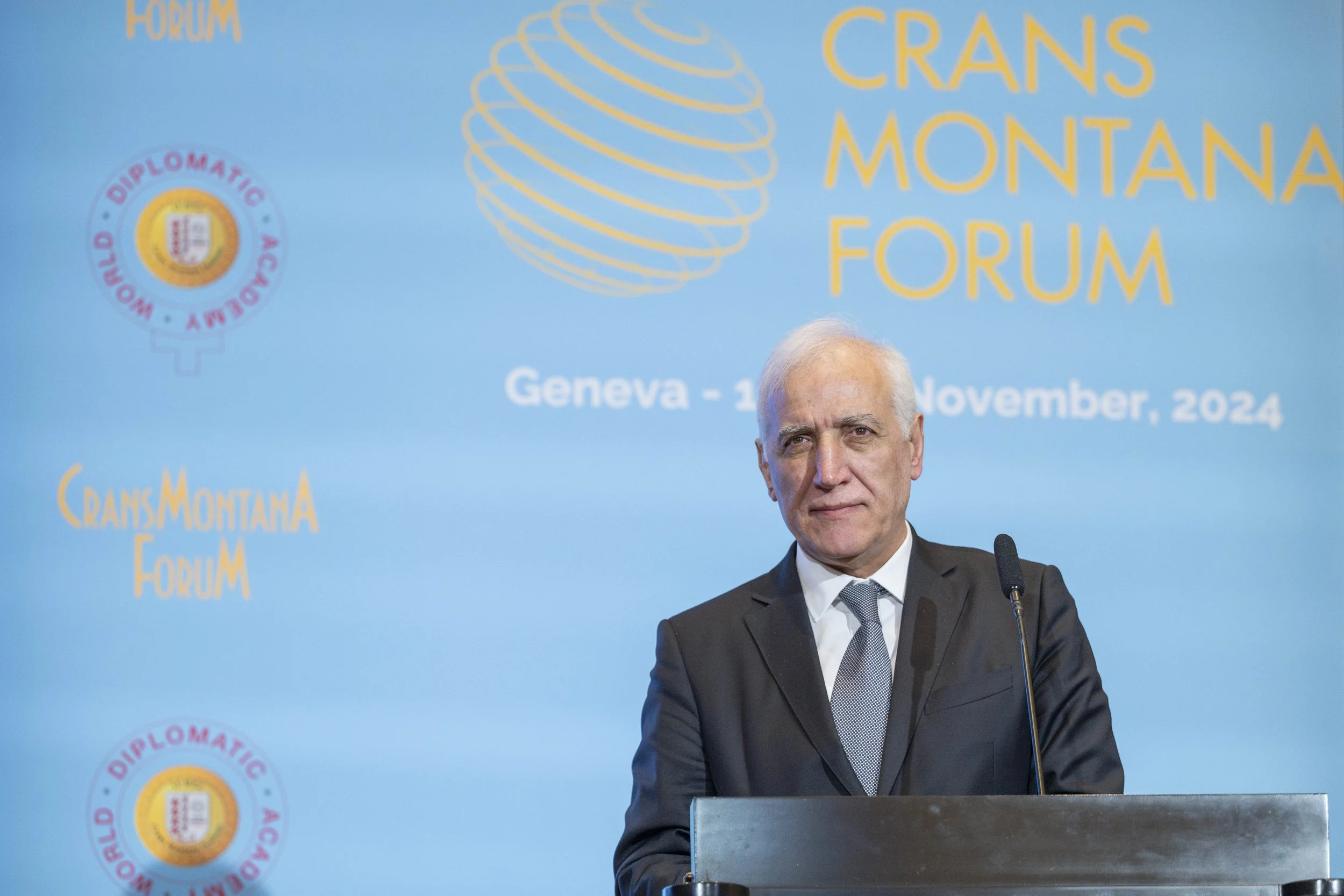 Un homme d'âge moyen portant un costume sombre, une chemise blanche et une cravate, se tient derrière un pupitre lors du forum Crans Montana à Genève en novembre 2024. Le fond est bleu avec un logo graphique et le texte du forum.