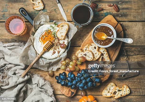 An rustic wooden table with a breakfast spread including a fried egg with bread, grapes, a jar of honey with a spoon, a glass of red wine, and slices of bread. There are also a knife and some torn bread pieces.