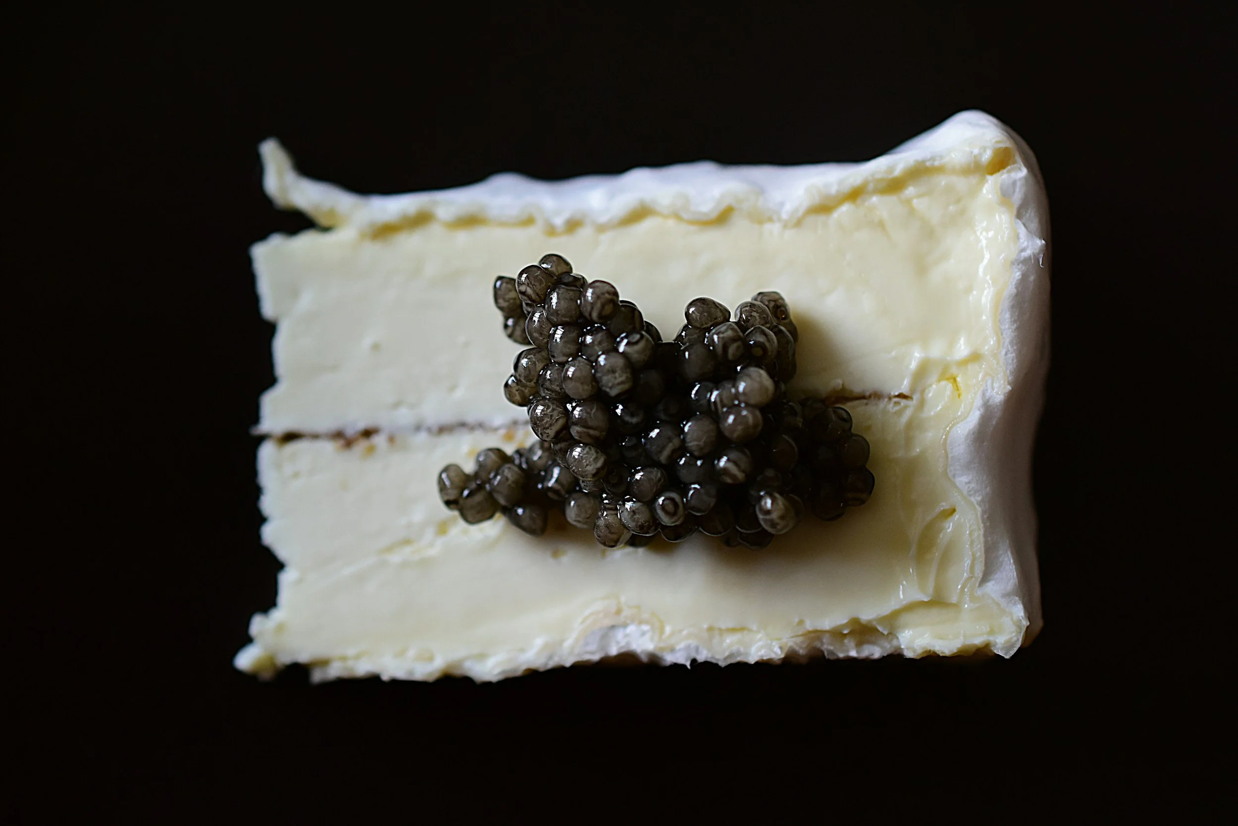 A wedge of soft cheese topped with black caviar, set against a black background.