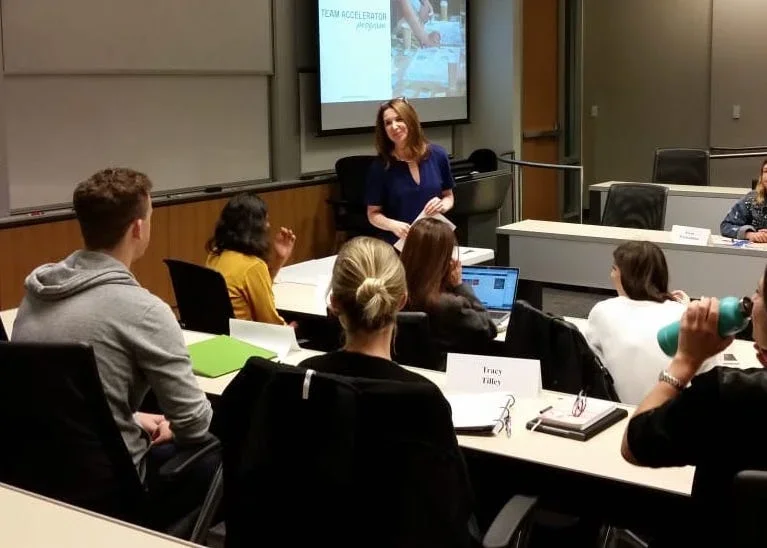 A classroom or conference room with a woman standing and presenting to a group of diverse adults seated around tables, with a presentation slide on the screen behind her.