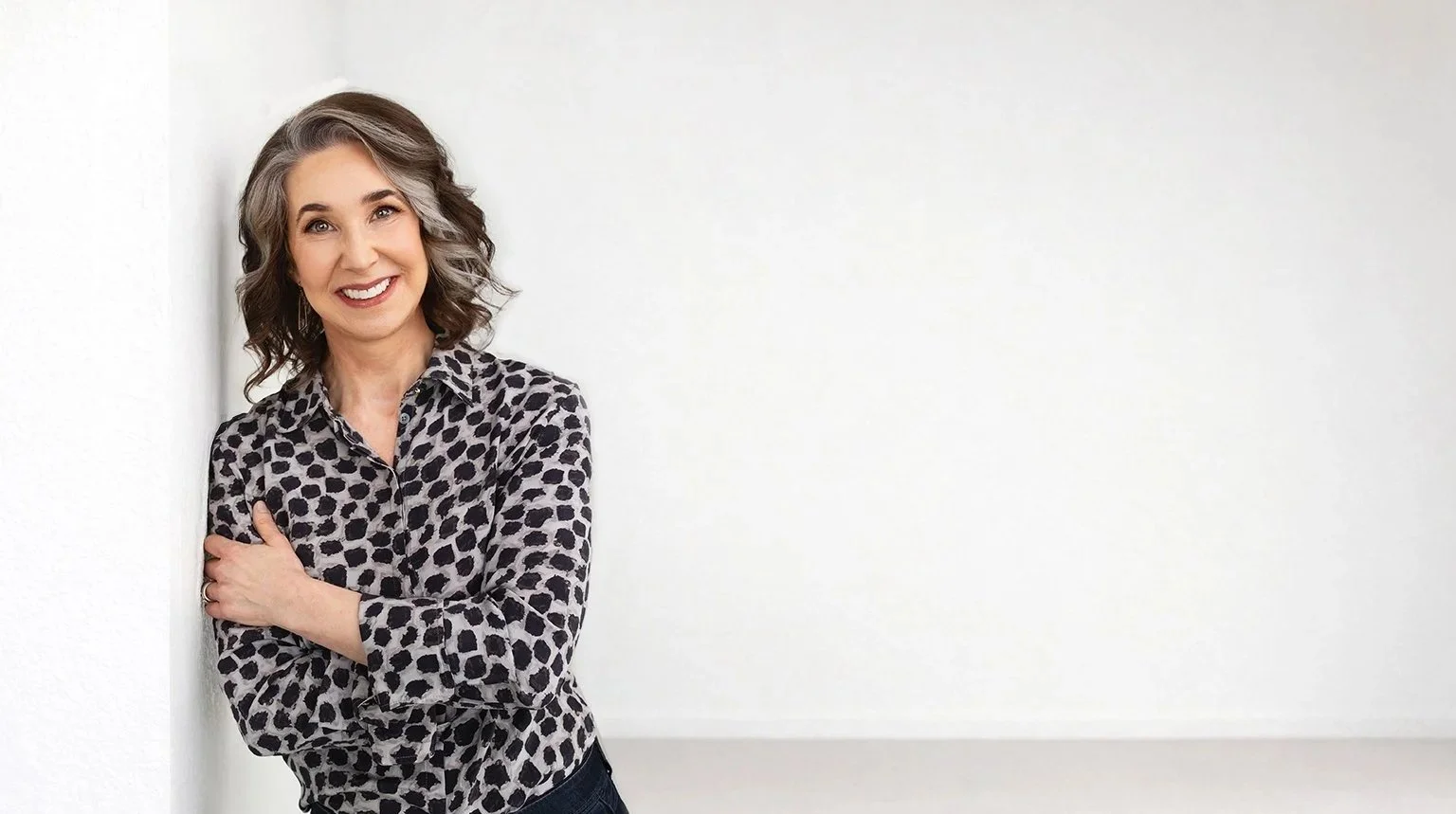 A woman with curly gray hair wearing a leopard print shirt, smiling and leaning against a white wall.