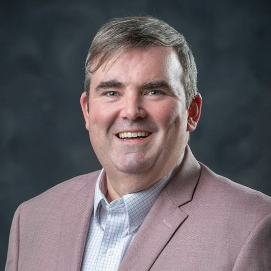 Professional headshot of a smiling man with light brown hair, wearing a light-colored blazer over a collared shirt, against a dark background.