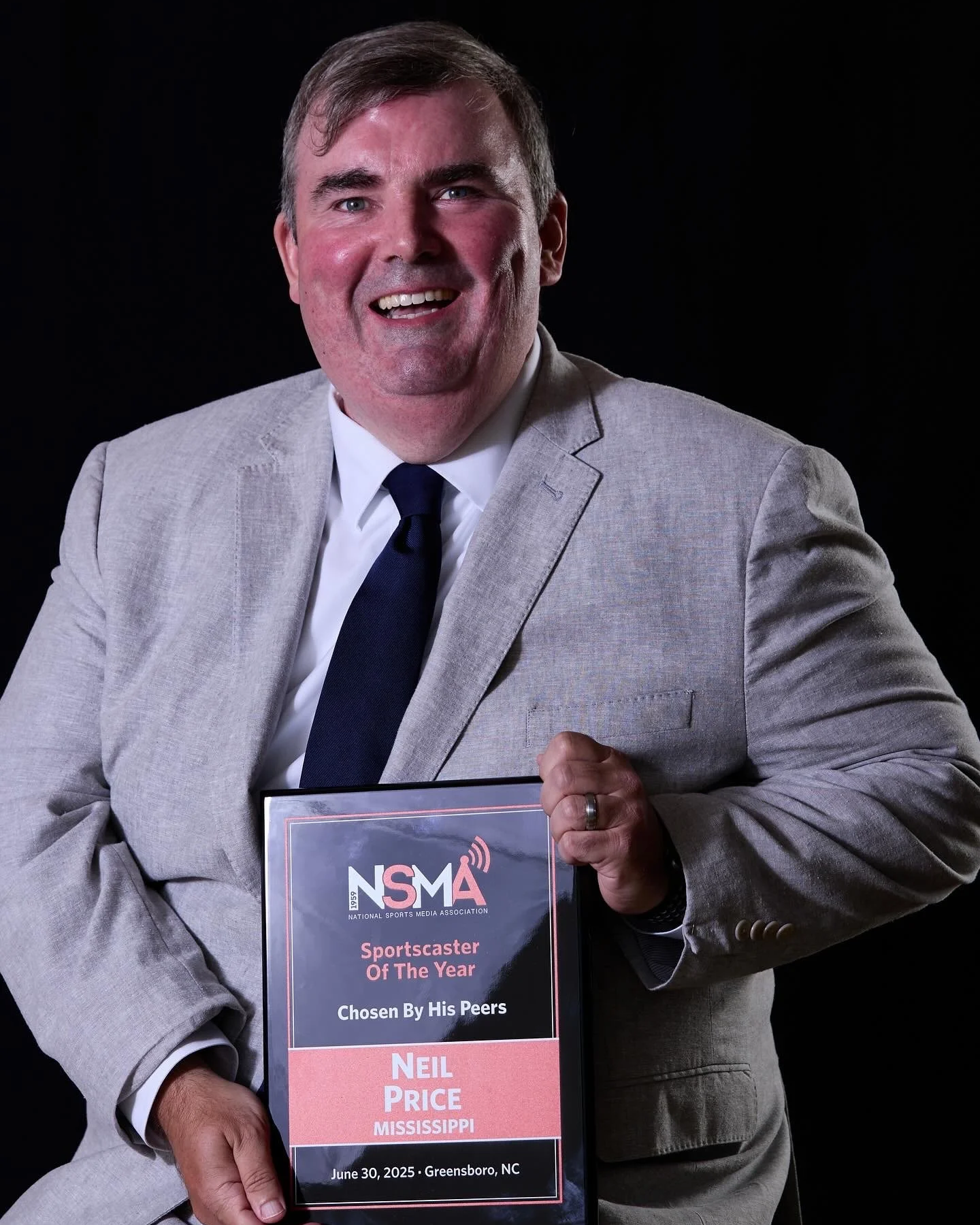 Man in a light gray suit holding an award plaque with the NSMA logo, text emphasizing Neil Price as Sportscaster of the Year, with a dark background.