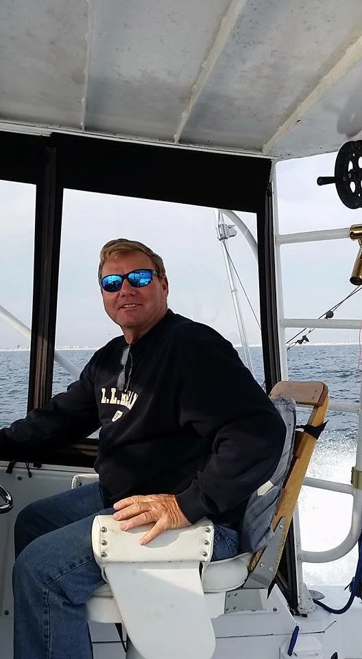 A man with sunglasses and a black jacket sitting on a boat with water in the background.