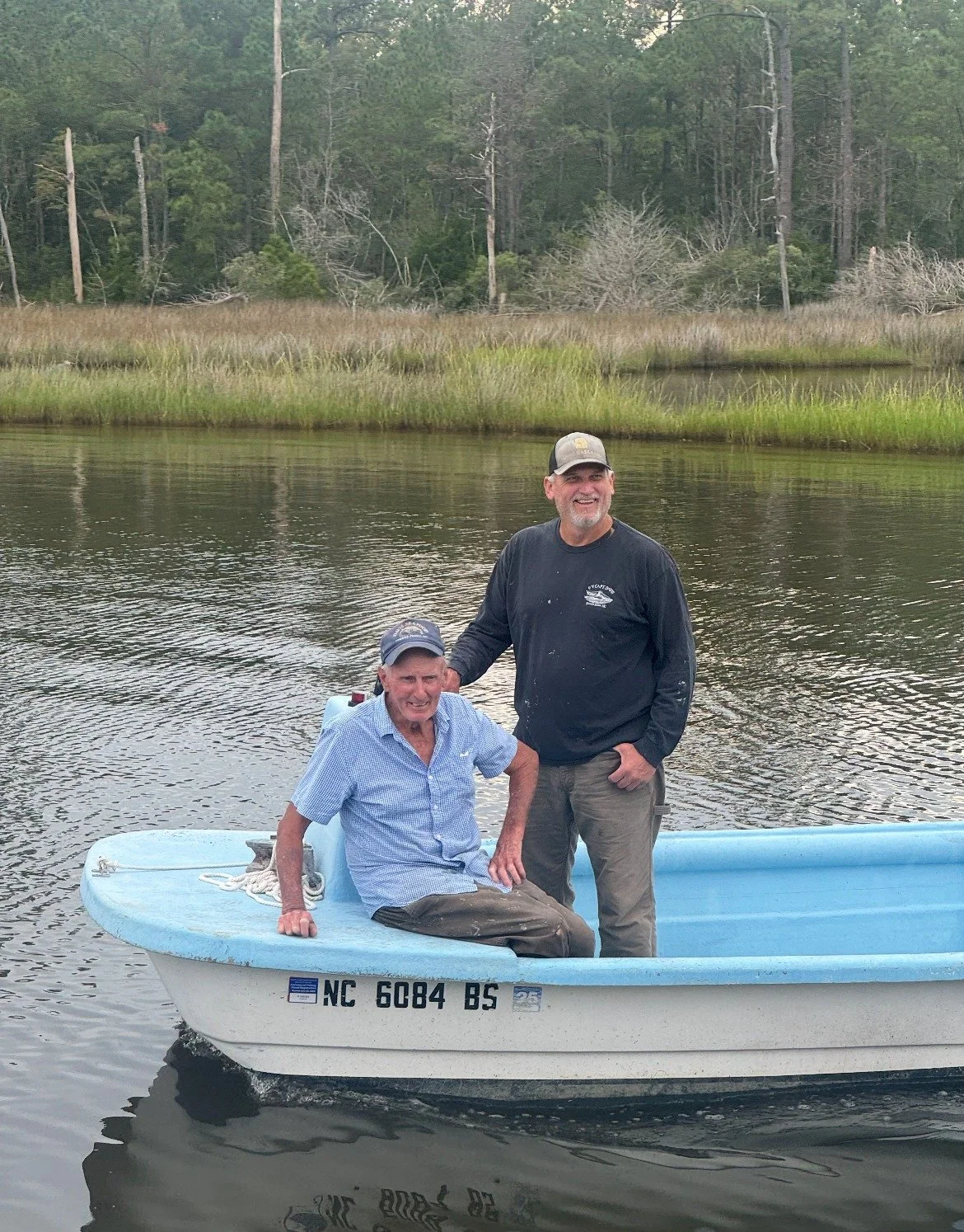 Two men in a small blue boat on a calm river, with a wooded shoreline in the background. One man is sitting and the other is standing, both smiling.