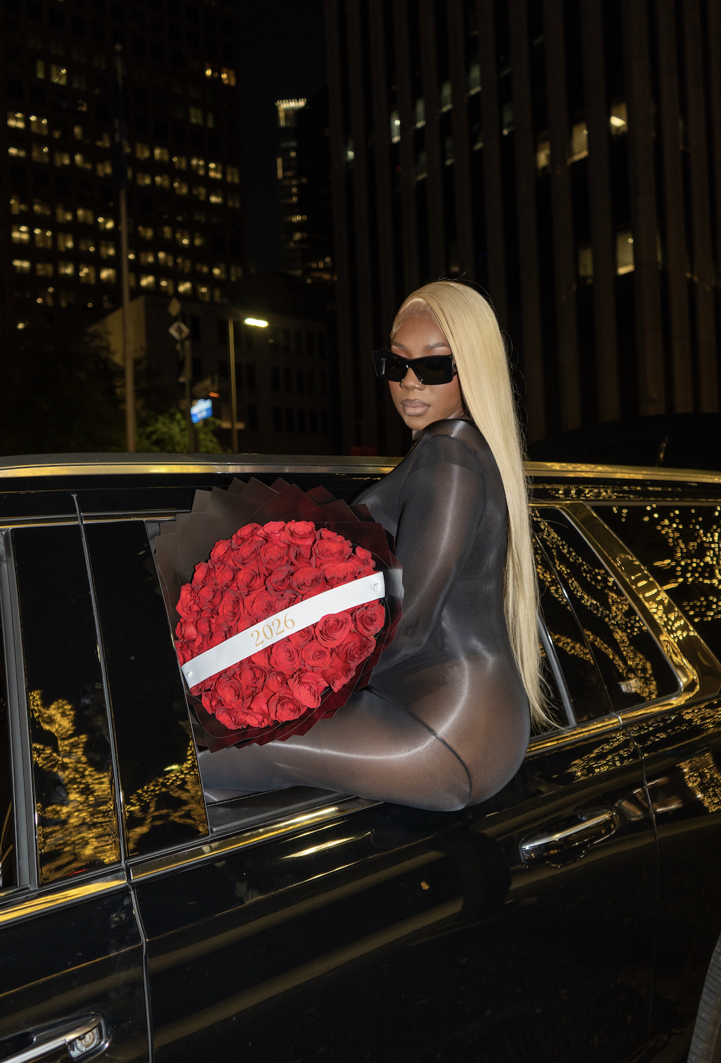 A woman with long blonde hair wearing black sunglasses, a black sheer outfit, sitting on the back of a black luxury car at night. She is holding a large bouquet of red roses with a white ribbon marked '2026'. City buildings with illuminated windows are visible in the background.