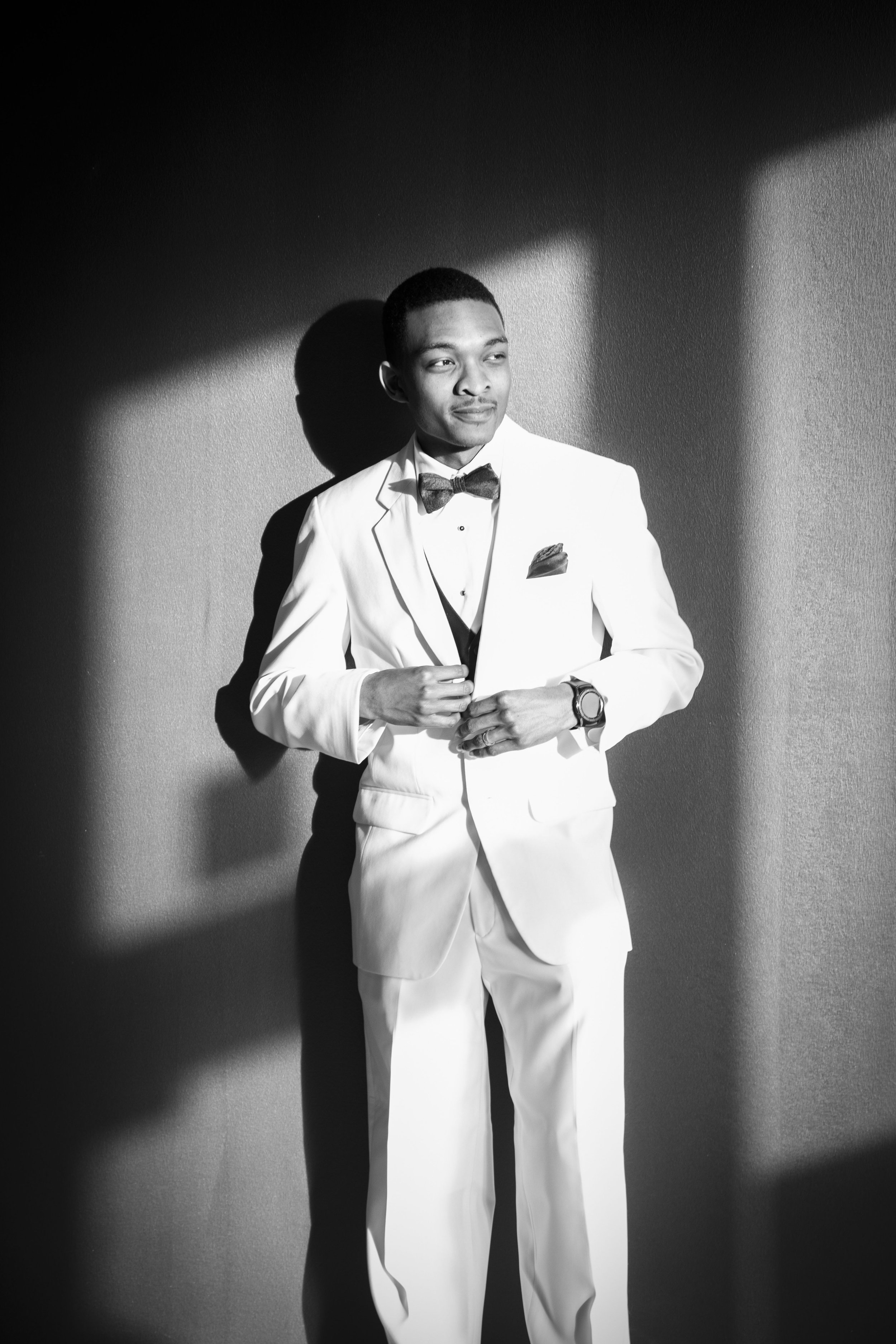 A young man in a white tuxedo with a bow tie, standing against a wall with shadows, looking away from the camera.
