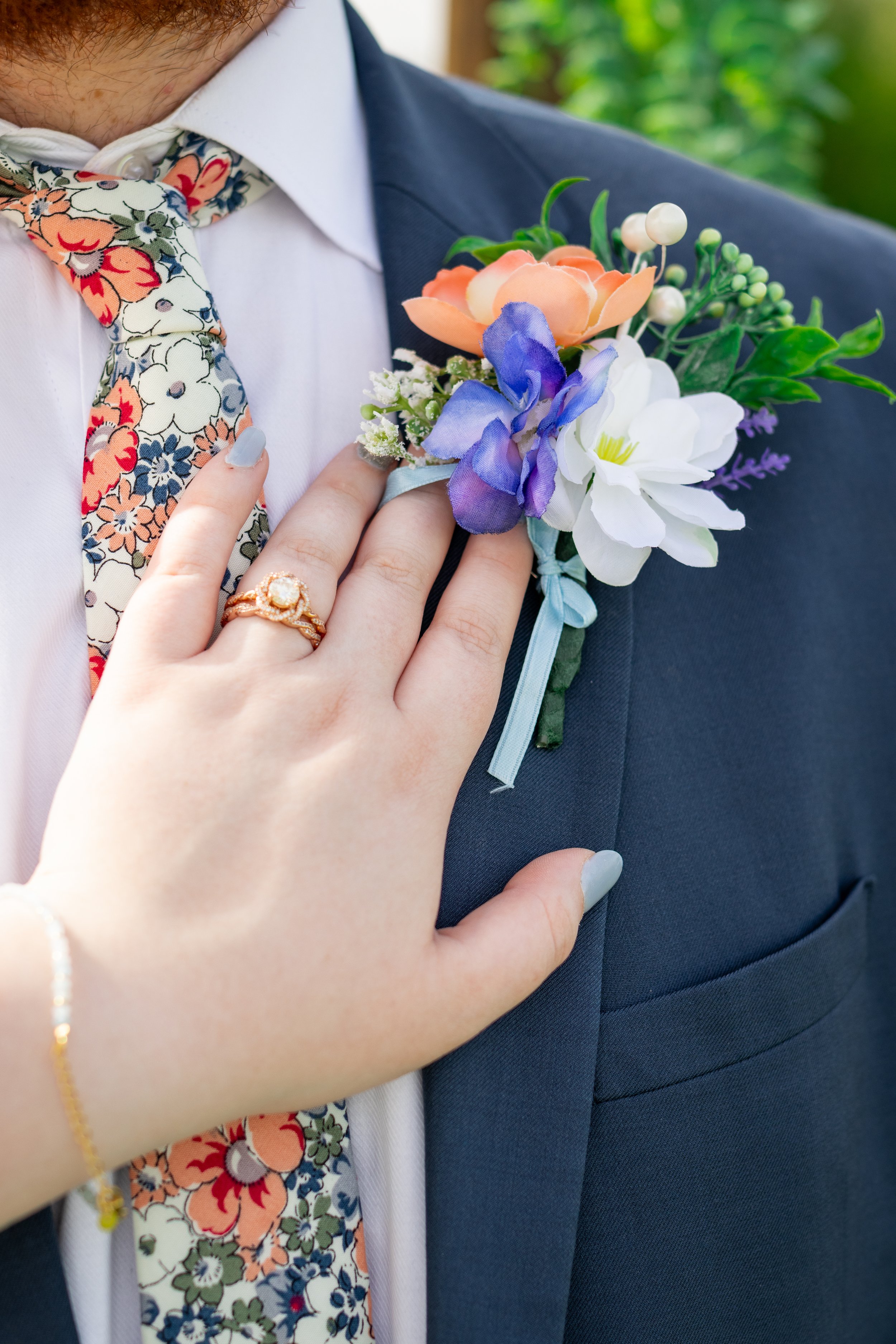 Close-up of a groom wearing a navy suit and a floral bow tie, with a colorful flower boutonniere on his lapel. A woman's hand with a wedding ring rests on his chest.
