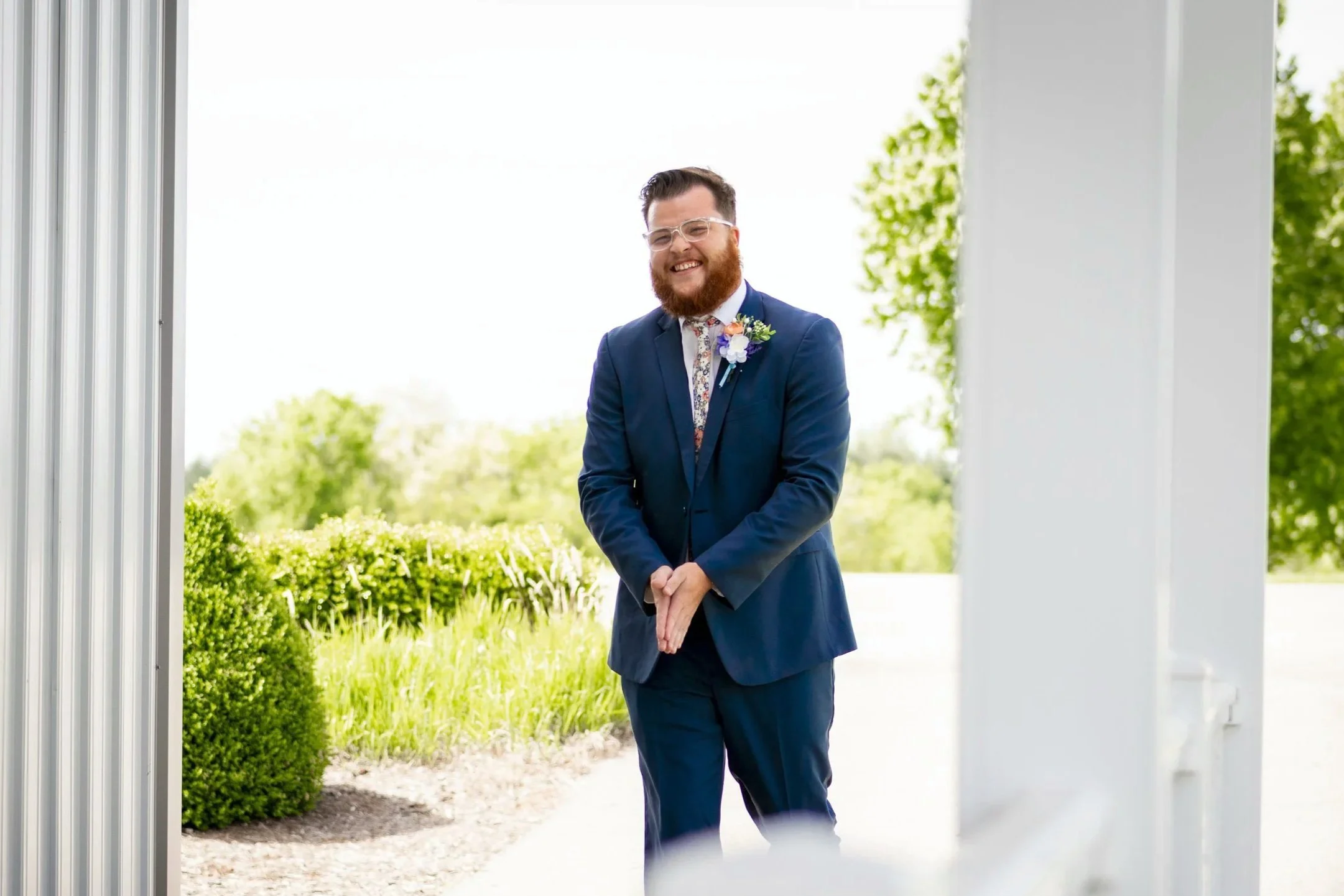 A smiling man in a dark blue suit with a boutonniere, standing outdoors in front of greenery, during a wedding ceremony.