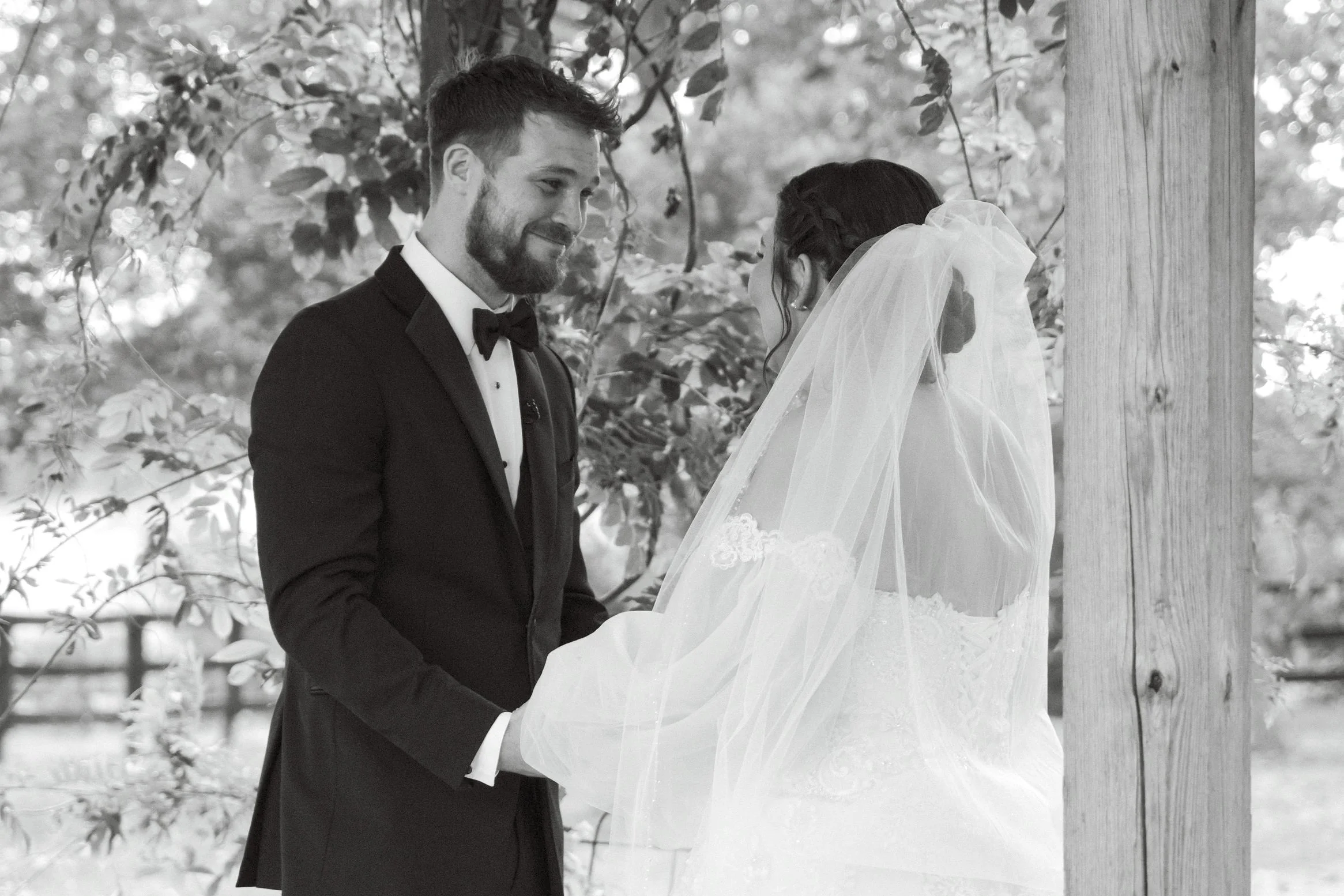 A black-and-white photo of a bride and groom during their wedding ceremony outdoors. The groom, in a tuxedo and bow tie, looks at the bride who is wearing a wedding dress and veil. They are holding hands and facing each other near a wooden post with 