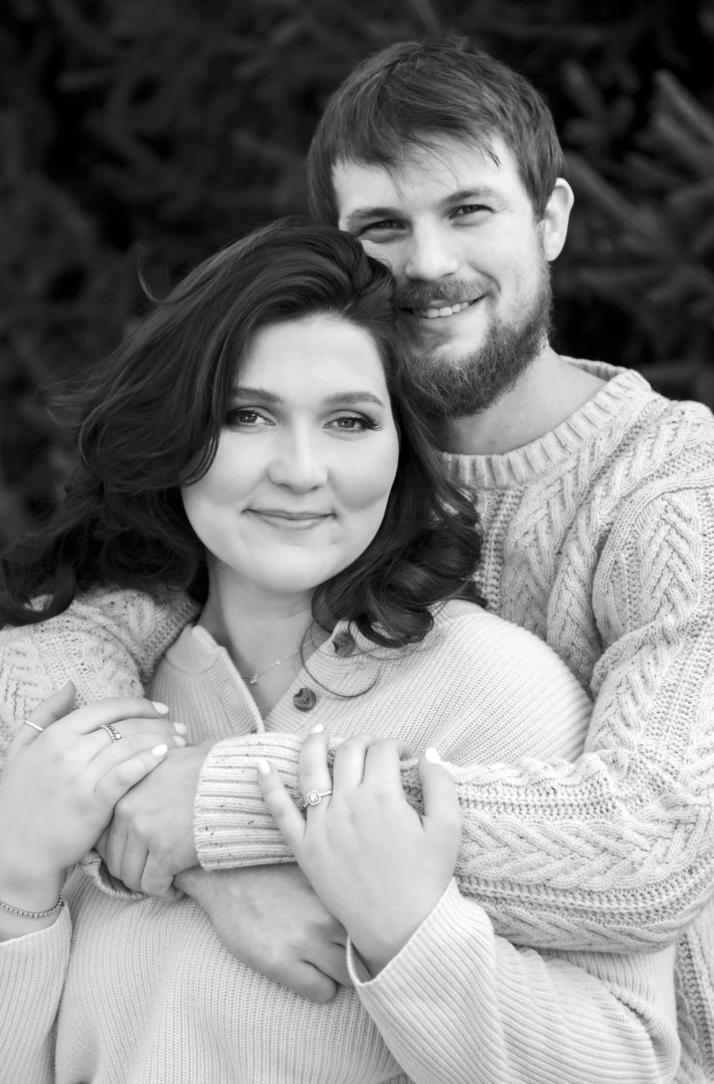A black and white photo of a smiling couple embracing each other outdoors. The woman has dark hair and is wearing a light-colored sweater, while the man has a beard and is wearing a textured knit sweater. They are holding hands with rings visible, an