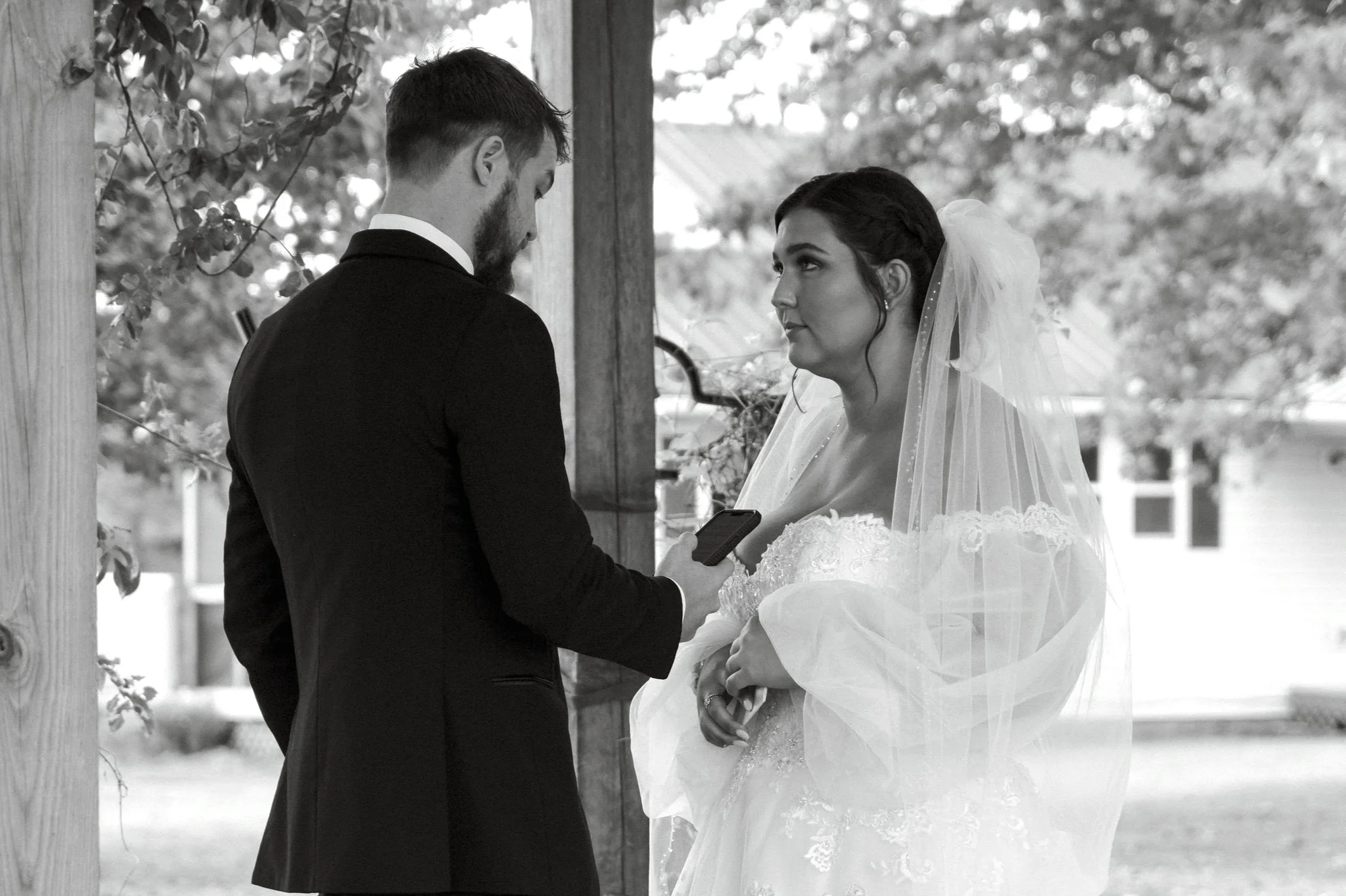 A bride and groom during their wedding ceremony, with the groom holding a phone and the bride wearing a wedding dress and veil, outdoors with trees in the background.
