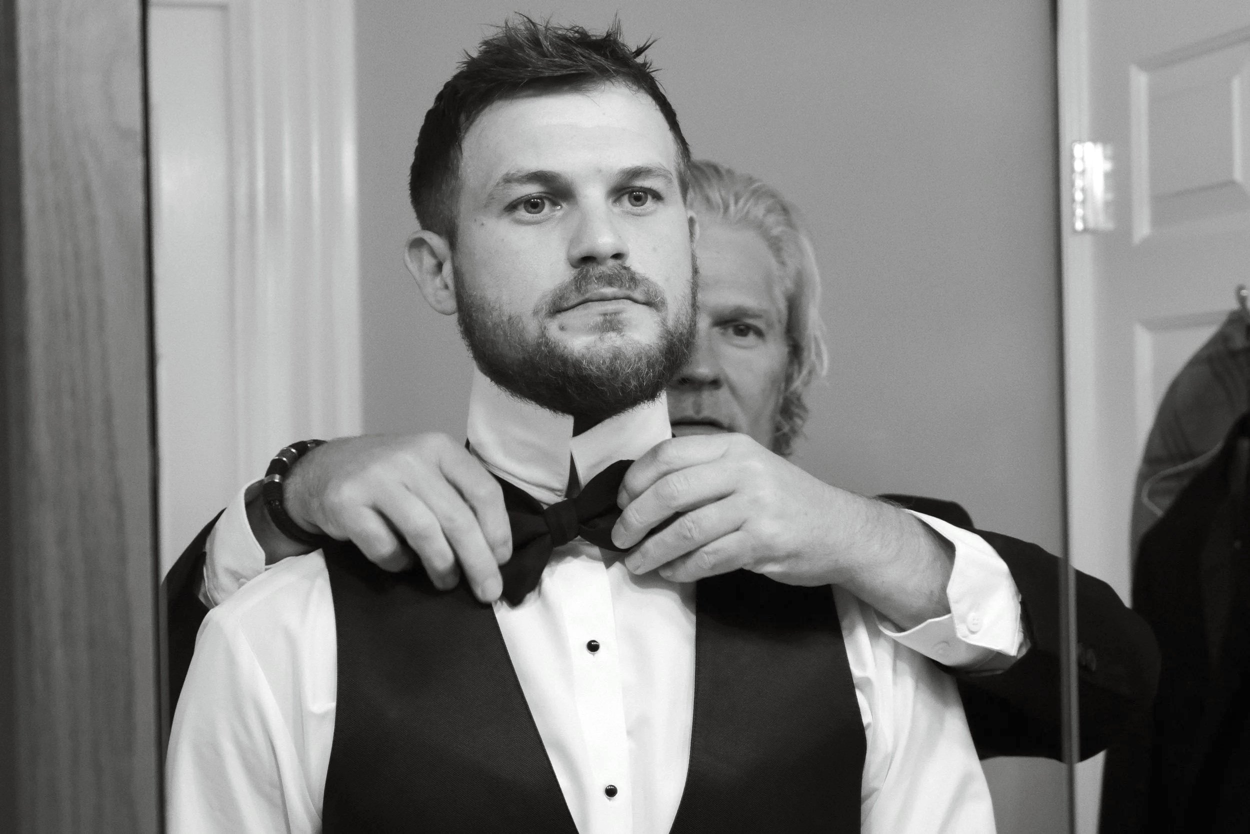 A man with a beard, wearing a tuxedo and bow tie, is having his bow tie adjusted by another person, reflected in a mirror.