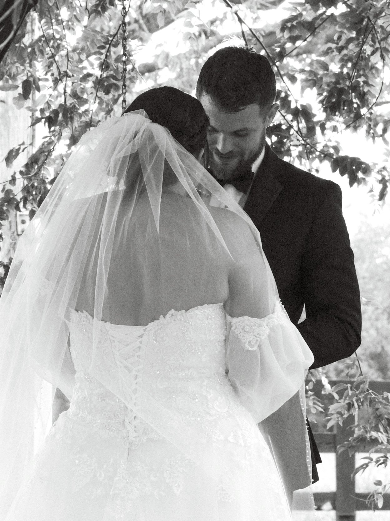 A black-and-white photo of a bride and groom in a close, intimate moment during their wedding, with the bride wearing a wedding gown and veil, and the groom in a suit, standing outdoors under a tree with leaves in the background.