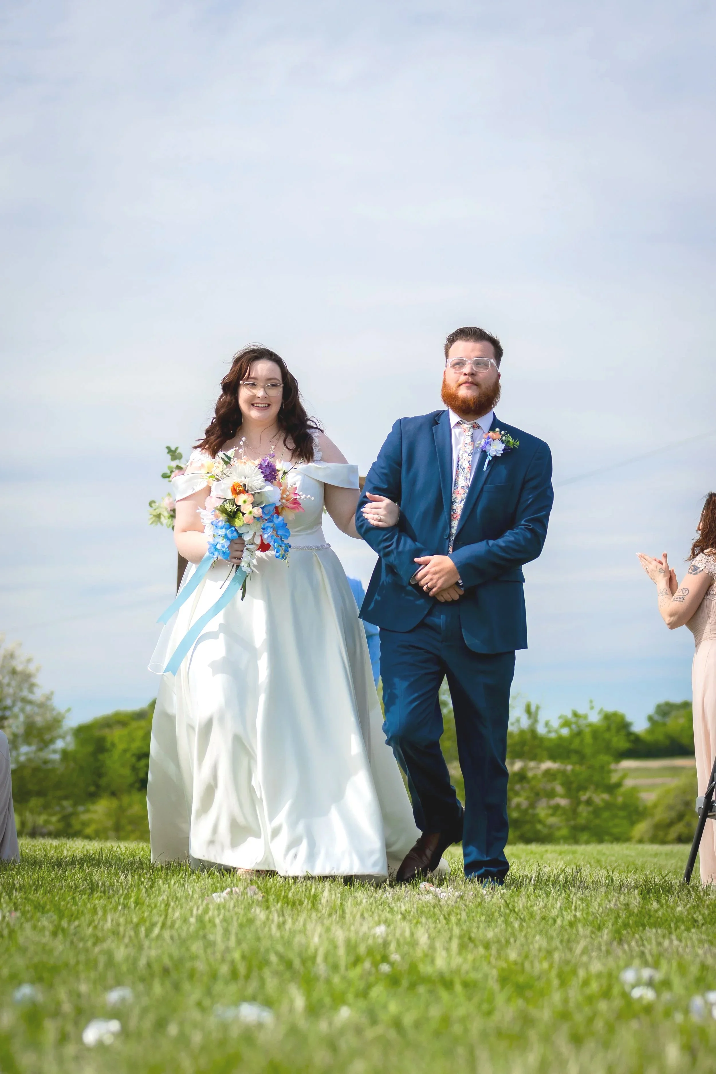 A bride in a white wedding dress holding a colorful bouquet walking arm-in-arm with a groom in a blue suit during an outdoor wedding ceremony on a grassy field.