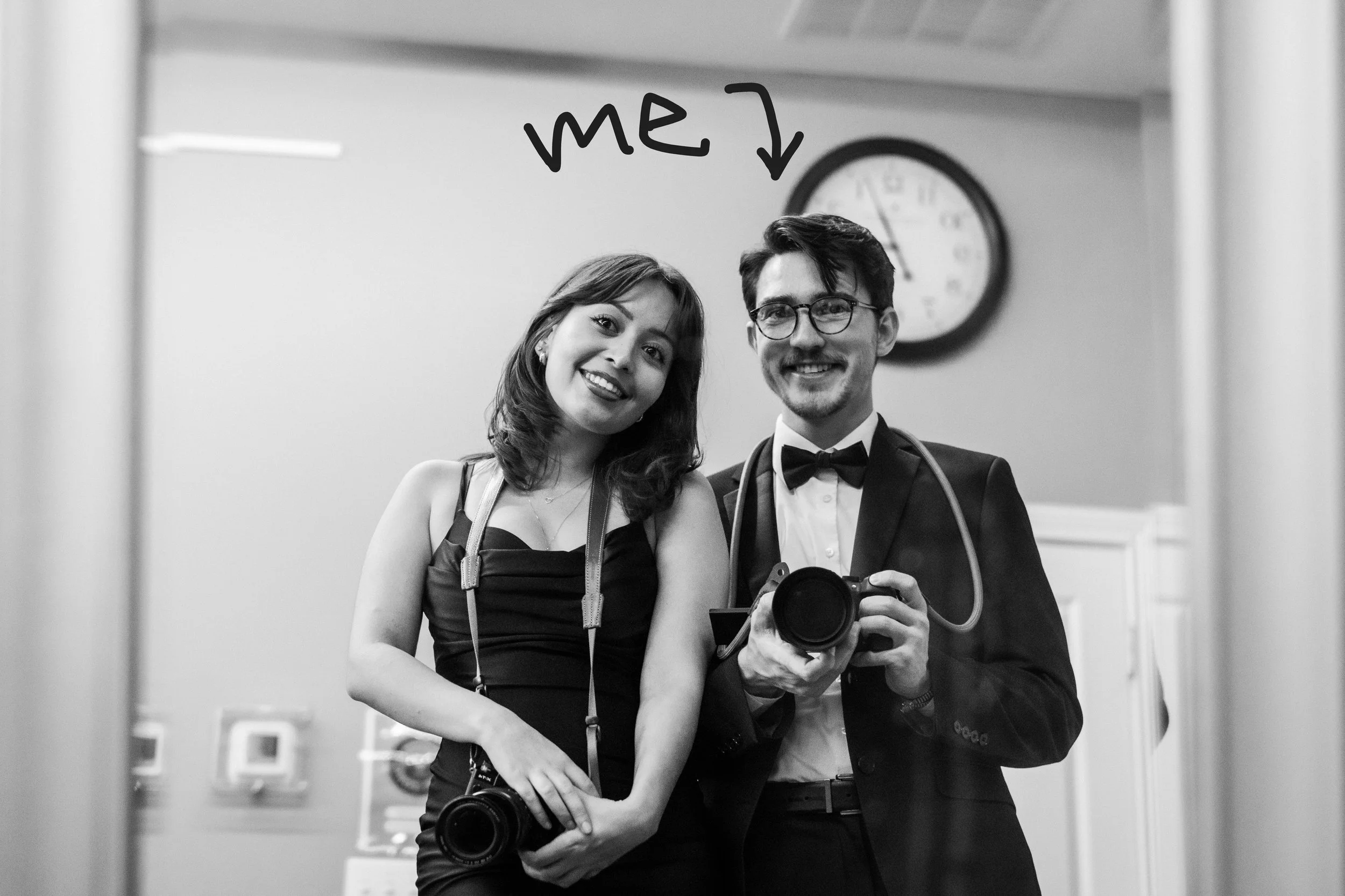 A young woman and a young man in formal attire taking a selfie through a mirror, with a handwritten note saying 'me' and an arrow pointing to the woman. They are smiling, and the man is holding a camera. There is a clock on the wall in the background.