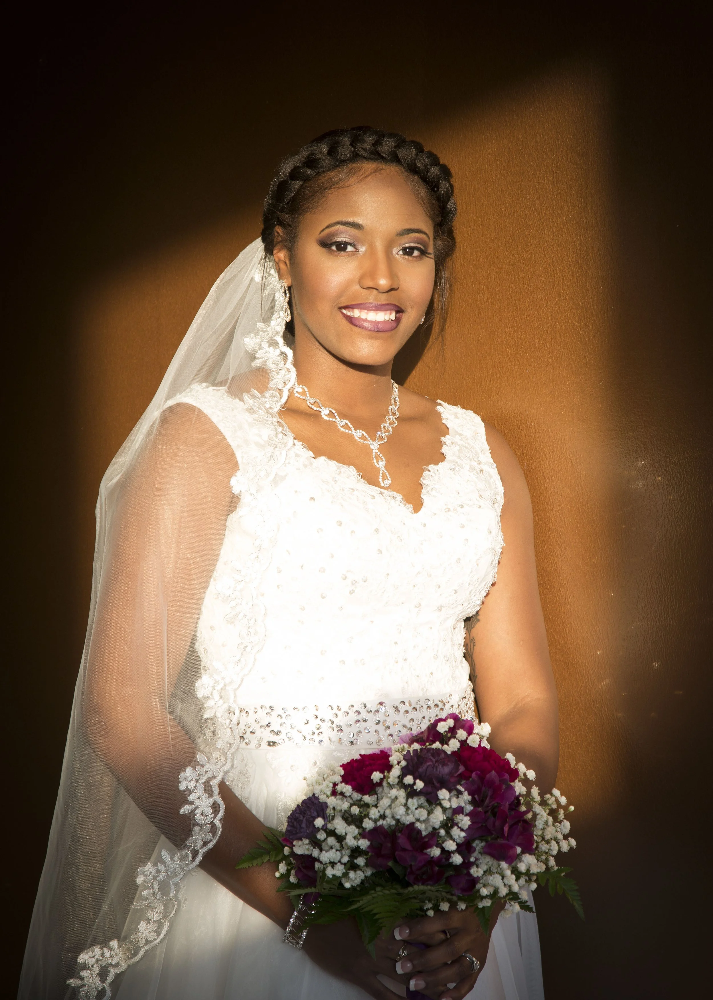 A bride wearing a white wedding dress with lace details, holding a bouquet of purple, white, and red flowers, standing against a brown background.