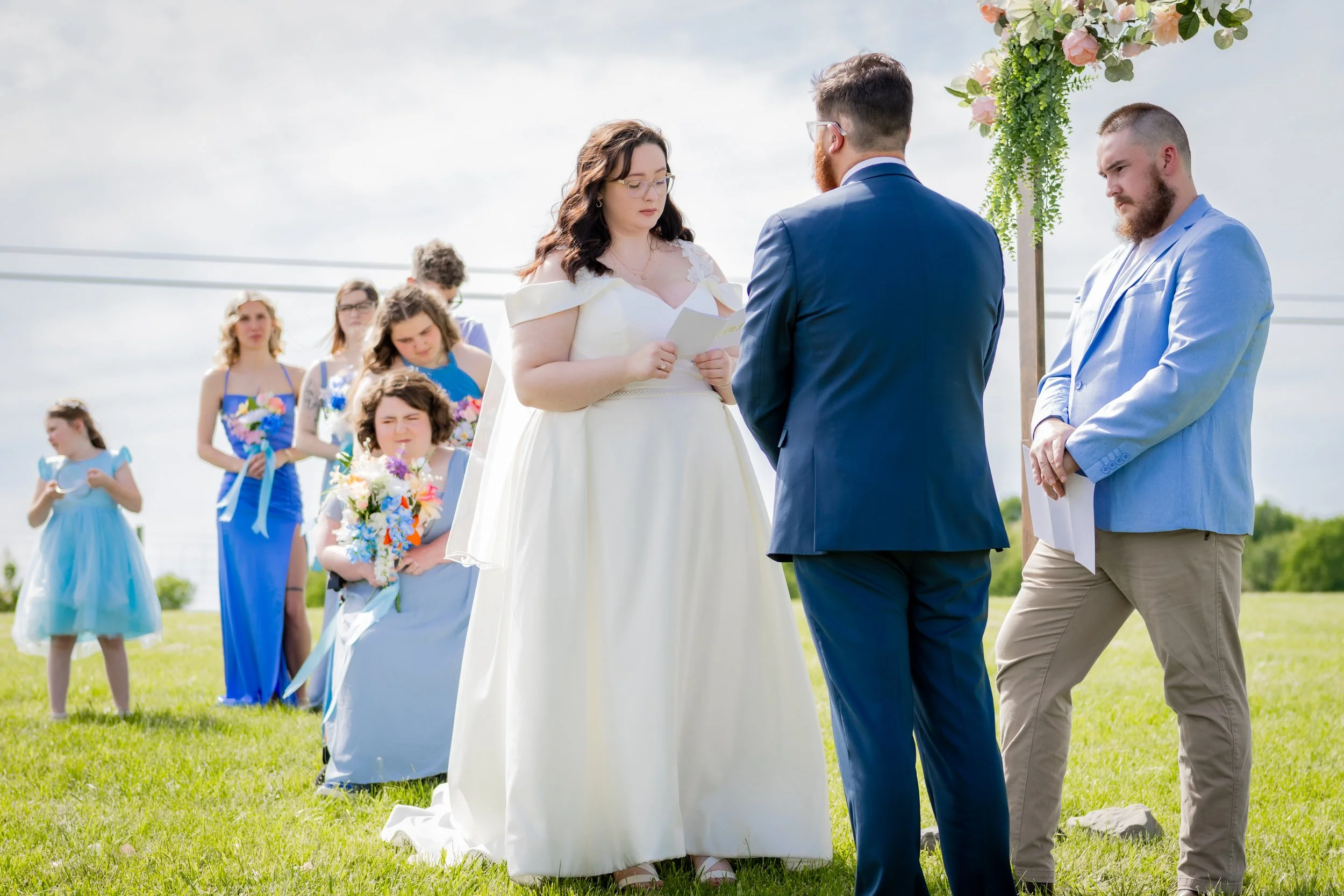 A couple is getting married outdoors in a grassy area, facing each other with the officiant in between them. The bride is wearing a white wedding dress and reading from a paper, while the groom is in a blue suit. The officiant is in a light blue blaz