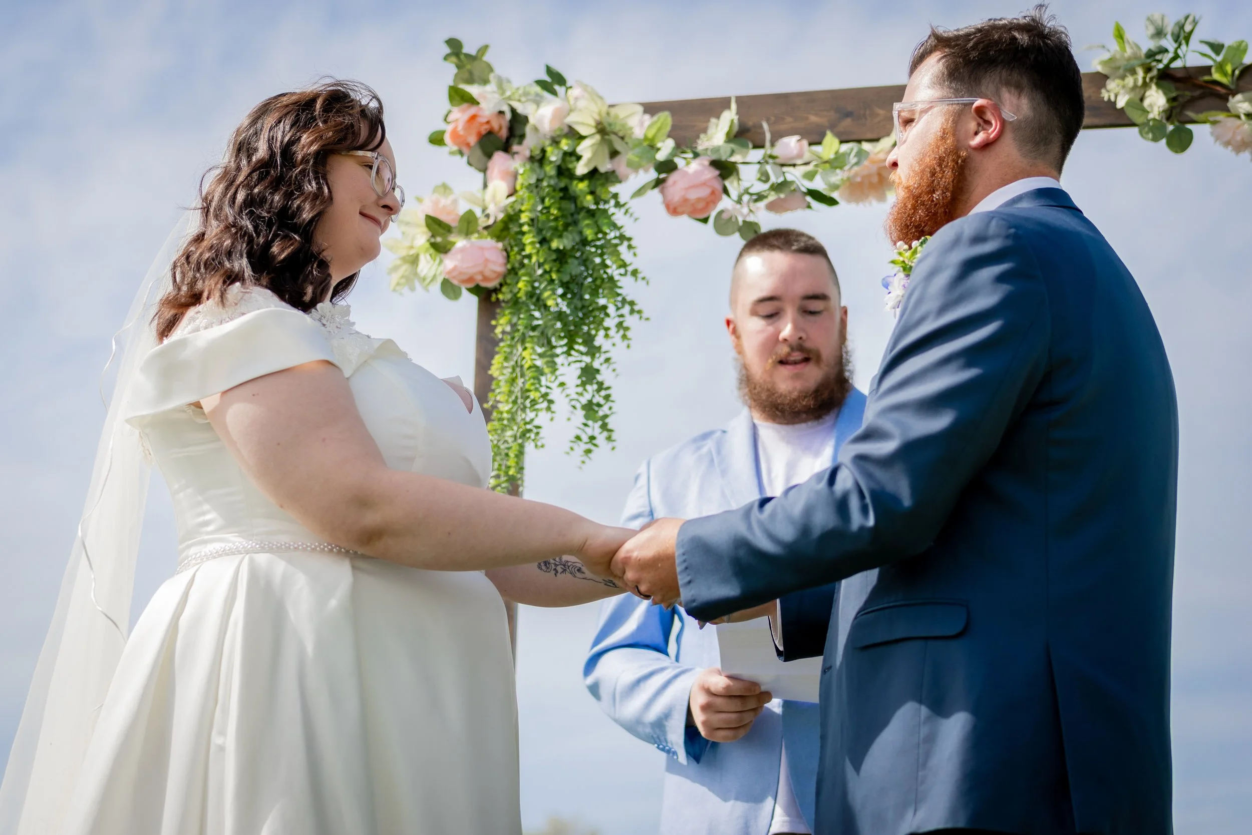 A couple gets married outdoors, holding hands and exchanging vows under a decorated wedding arch with pink and white flowers, as officiant reads during daytime.