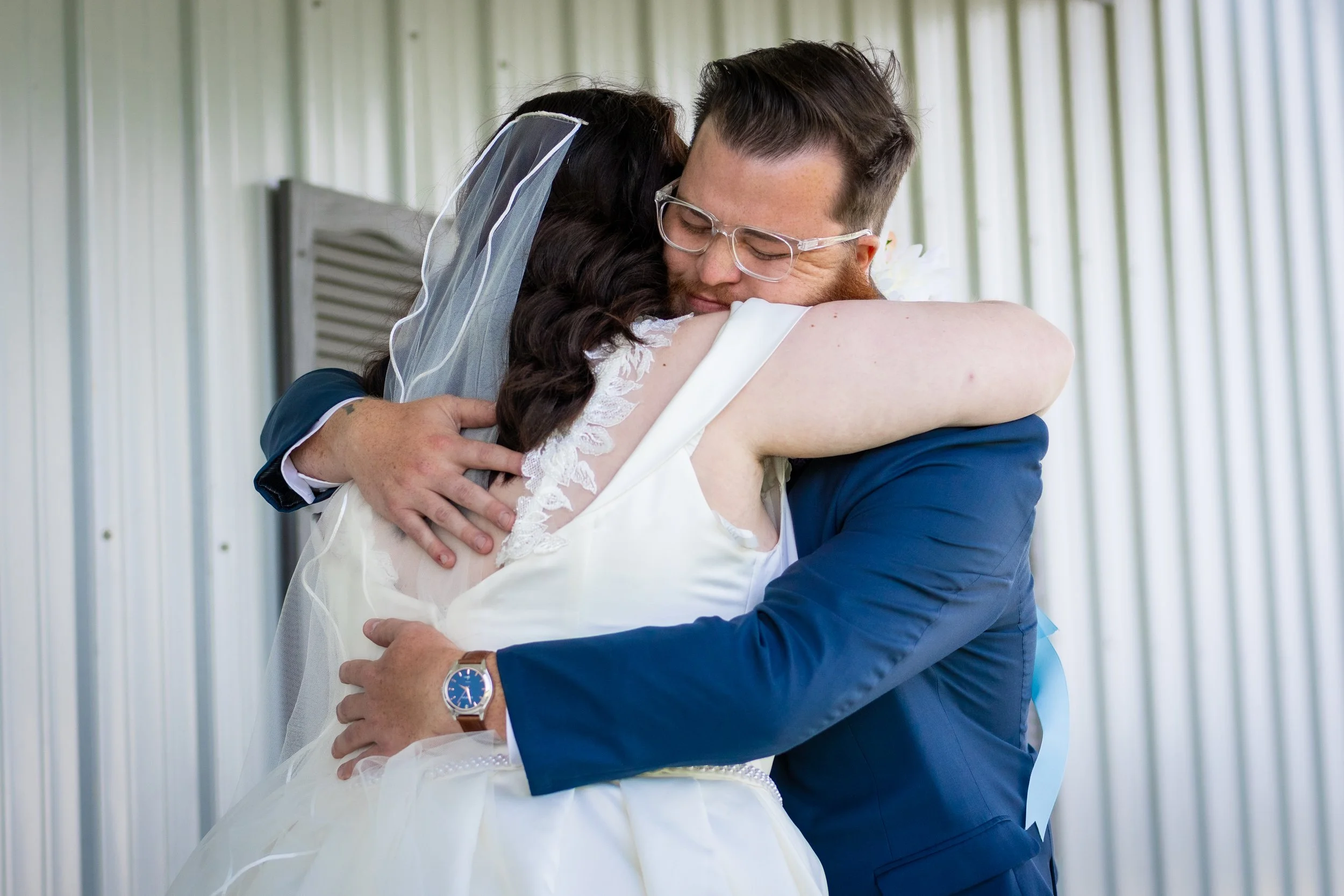 A couple hugging, with the bride in a white wedding dress and the groom in a blue suit, inside a room with metallic walls.
