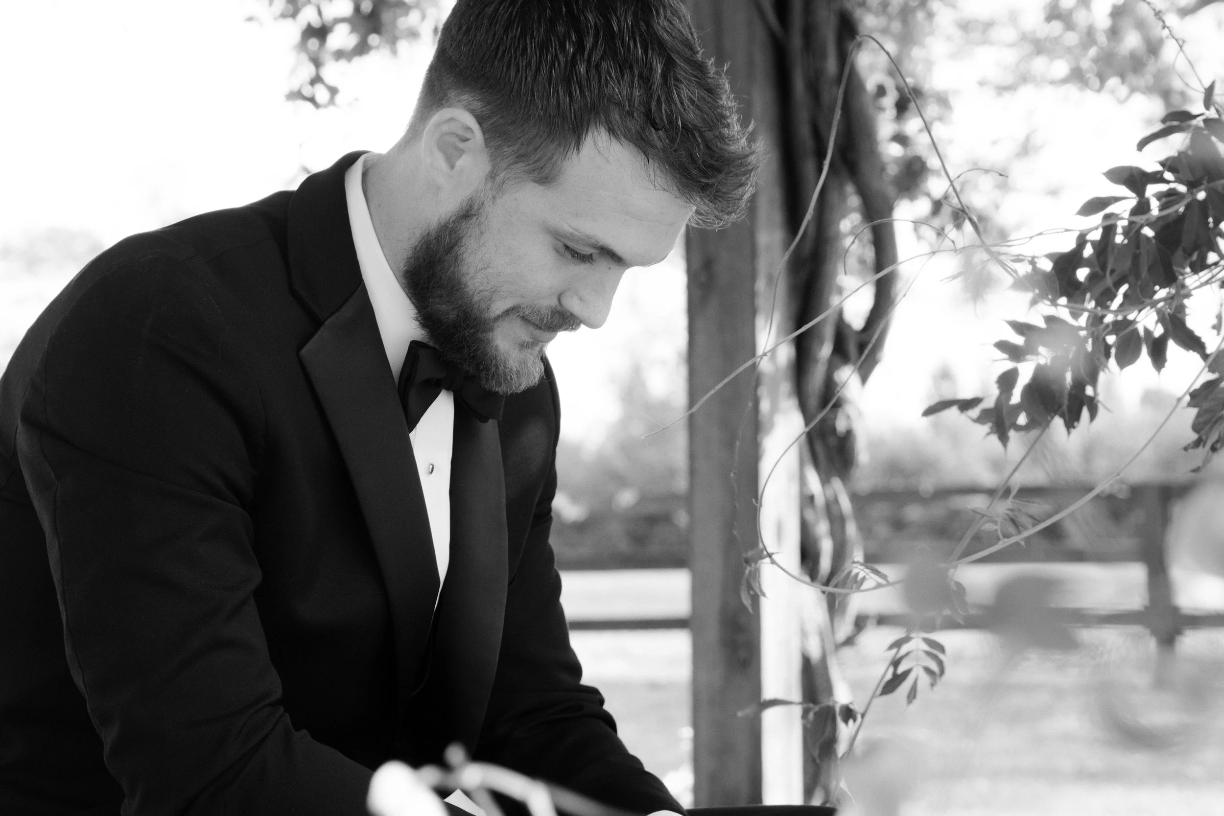 A man with a beard, dressed in formal wedding attire, is looking down while leaning forward outdoors.