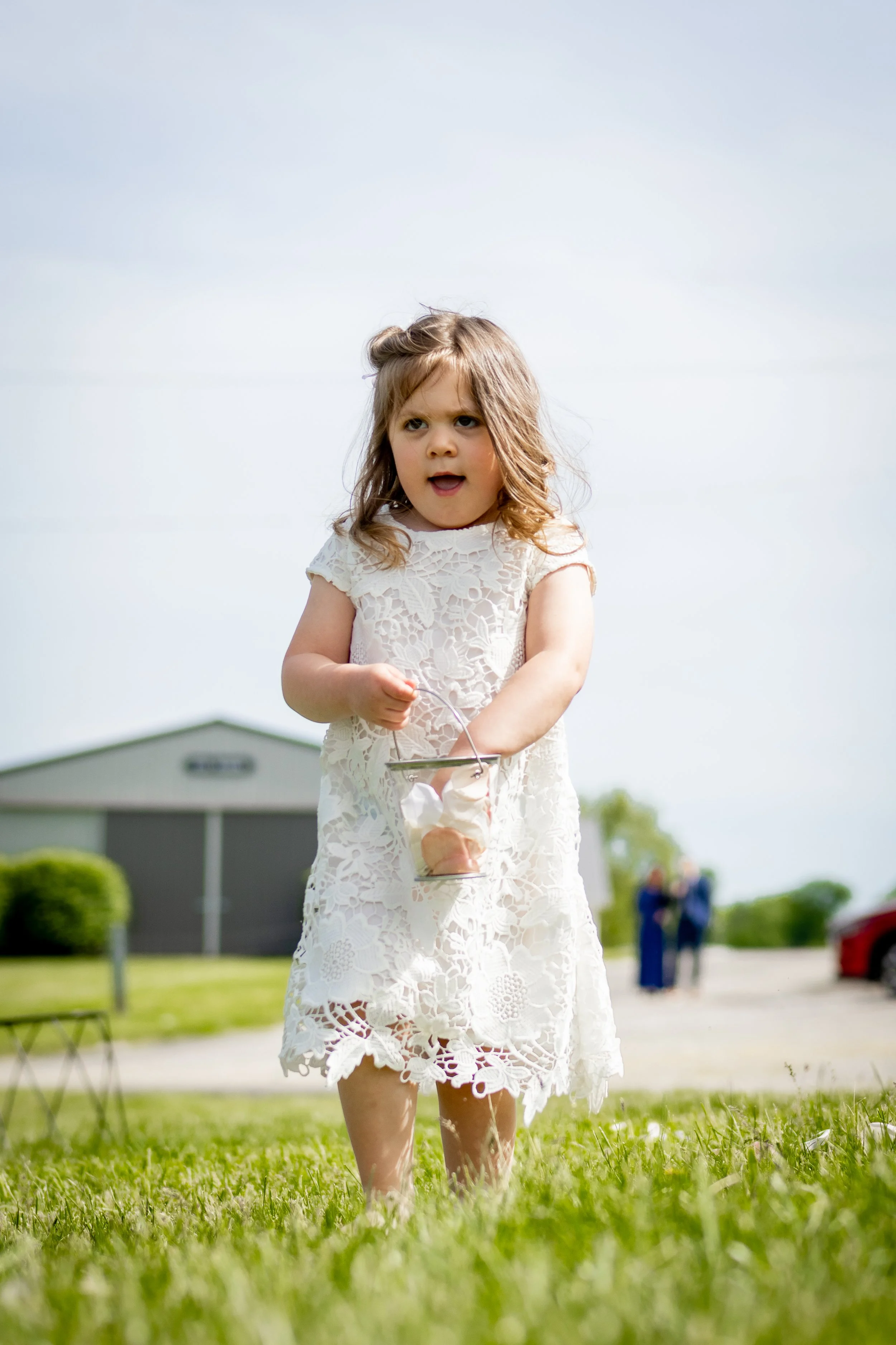 Young girl in a white lace dress holding a clear bucket with ice cream, standing outdoors on grass with a building and people in the background.