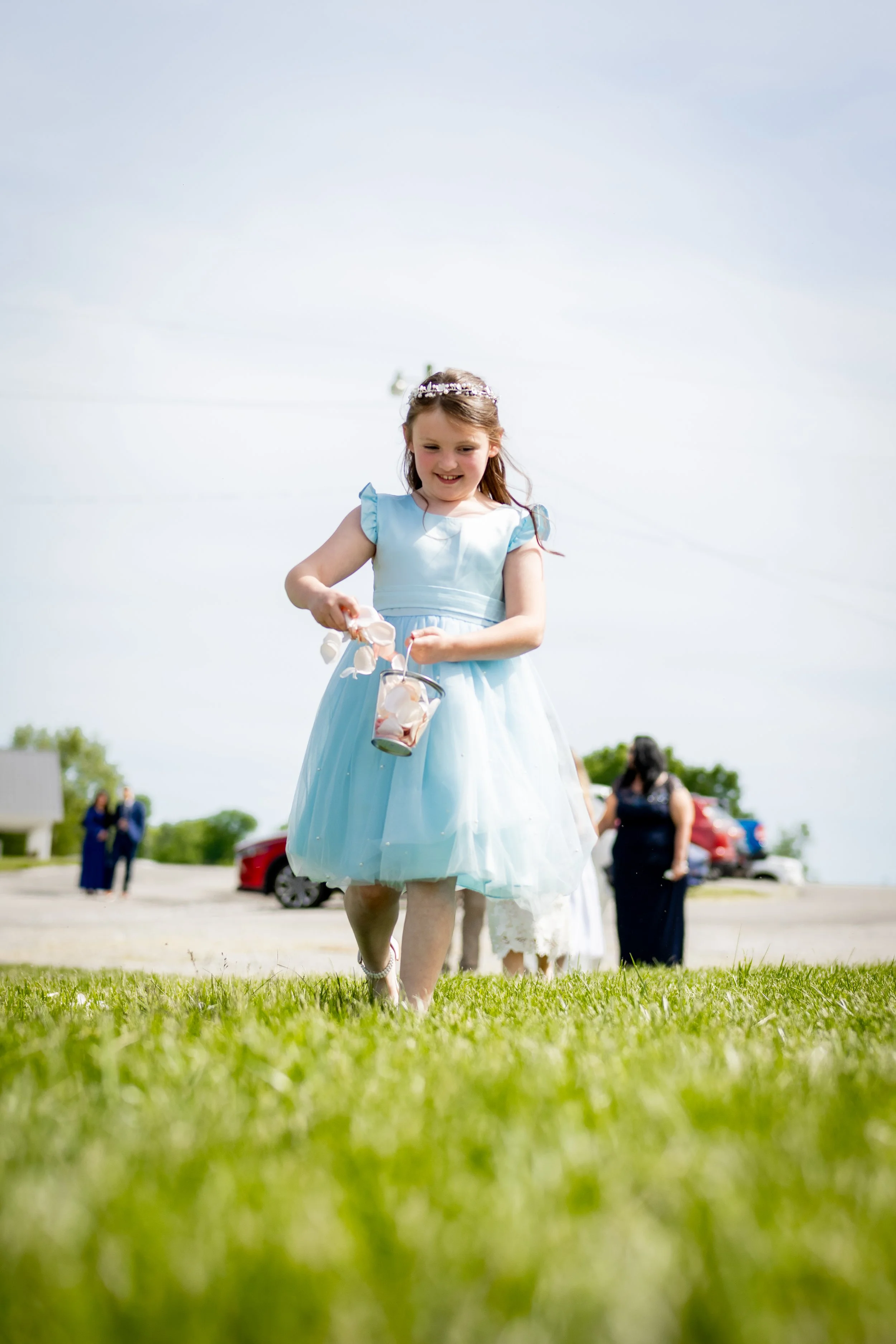A young girl in a light blue dress and tiara smiling as she walks on grass holding a small bucket and toy.