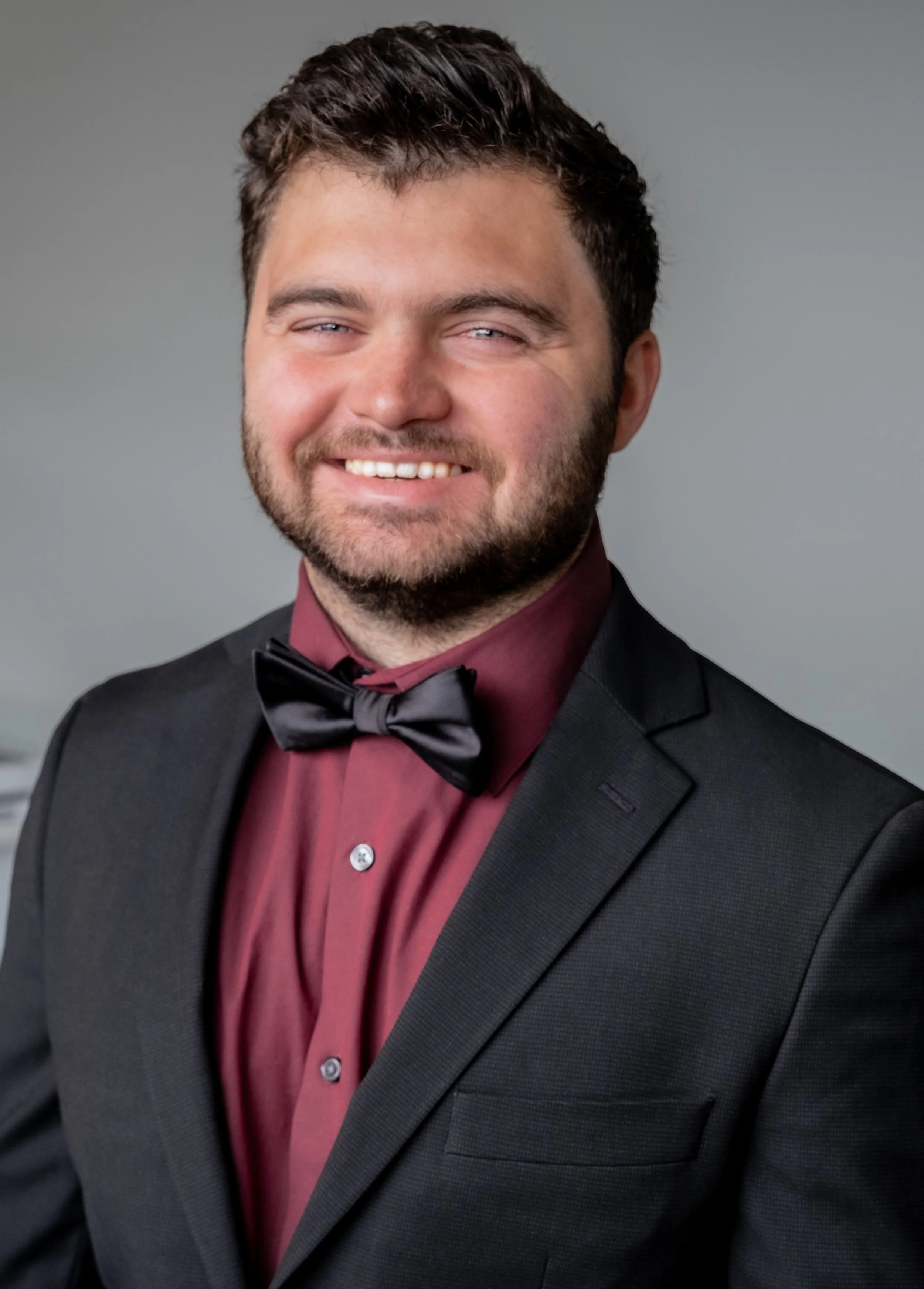 A man in a tuxedo with a red shirt and black bow tie, smiling against a grey background.