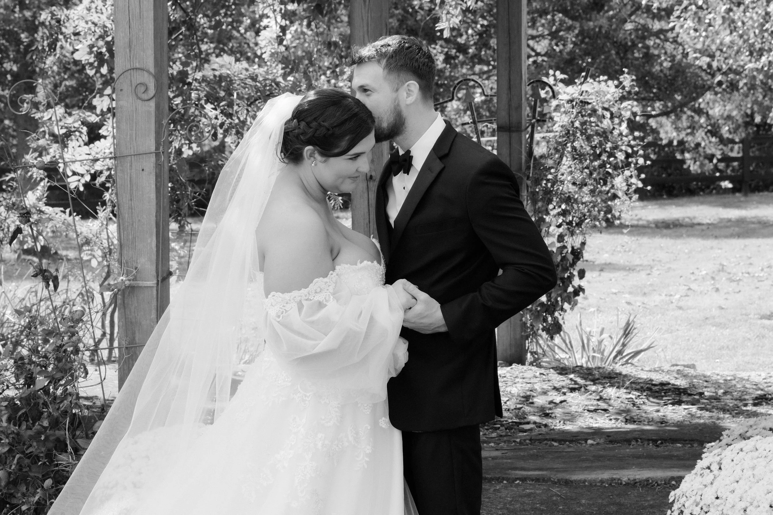 A black and white photo of a bride and groom holding hands, standing close with foreheads touching outdoors, with trees and a wooden fence in the background.