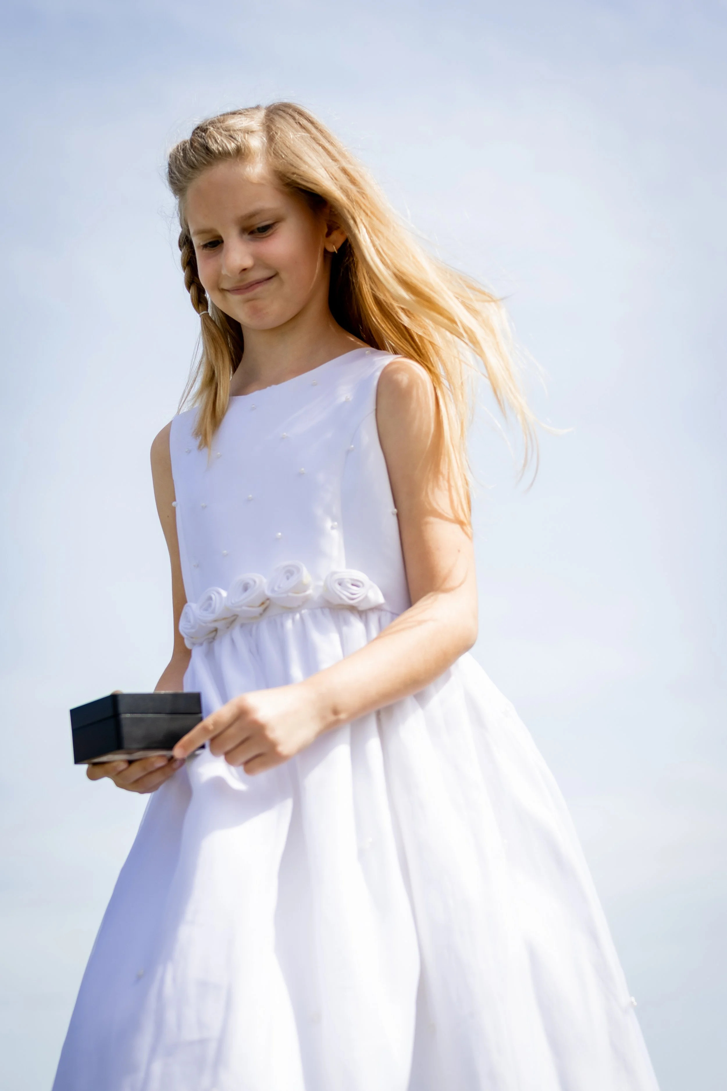 A young girl in a white dress holding a small black gift box, outdoors with a cloudy sky in the background.