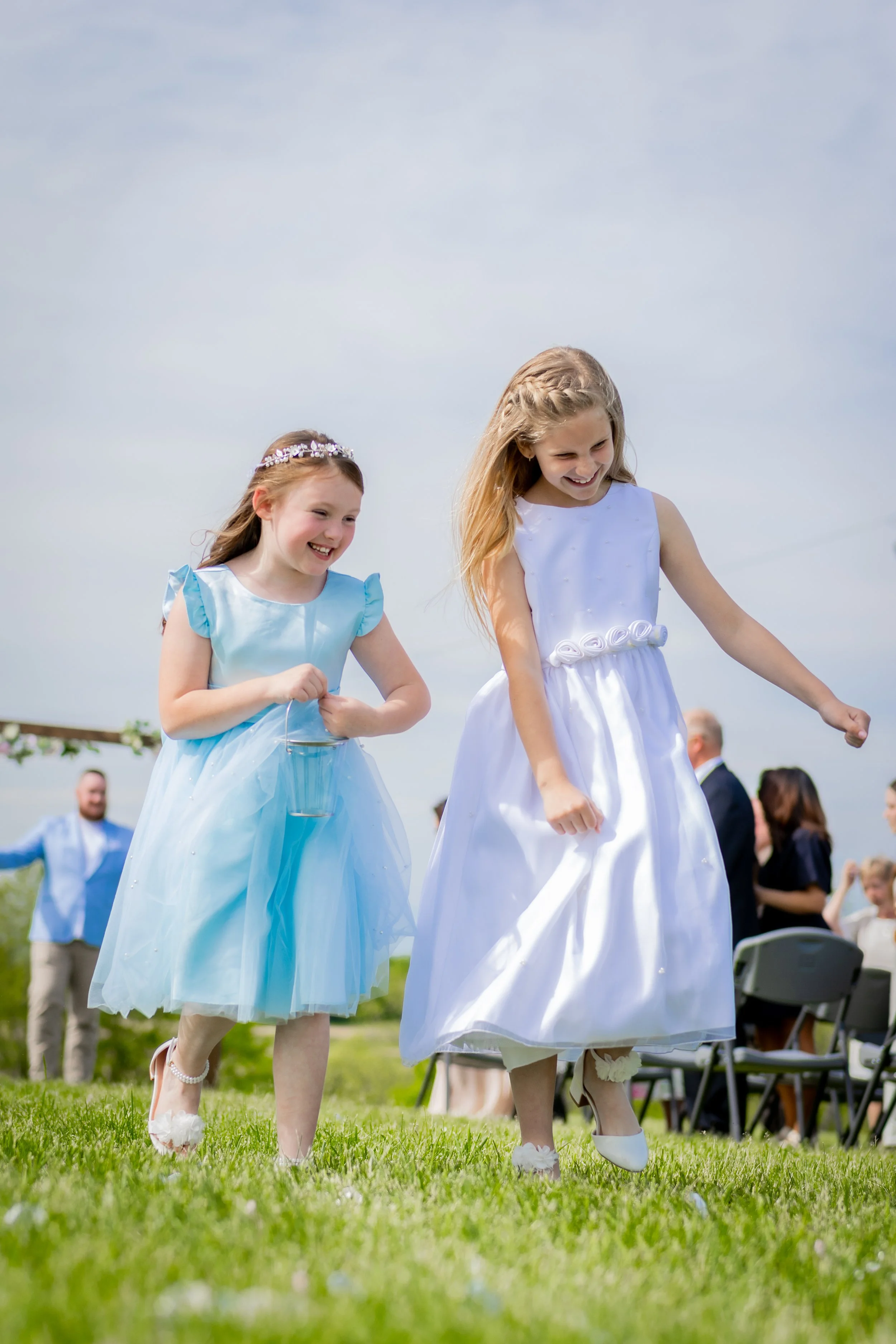 Two young girls in white and light blue dresses smiling and walking on grass at an outdoor wedding ceremony, with guests seated in the background.