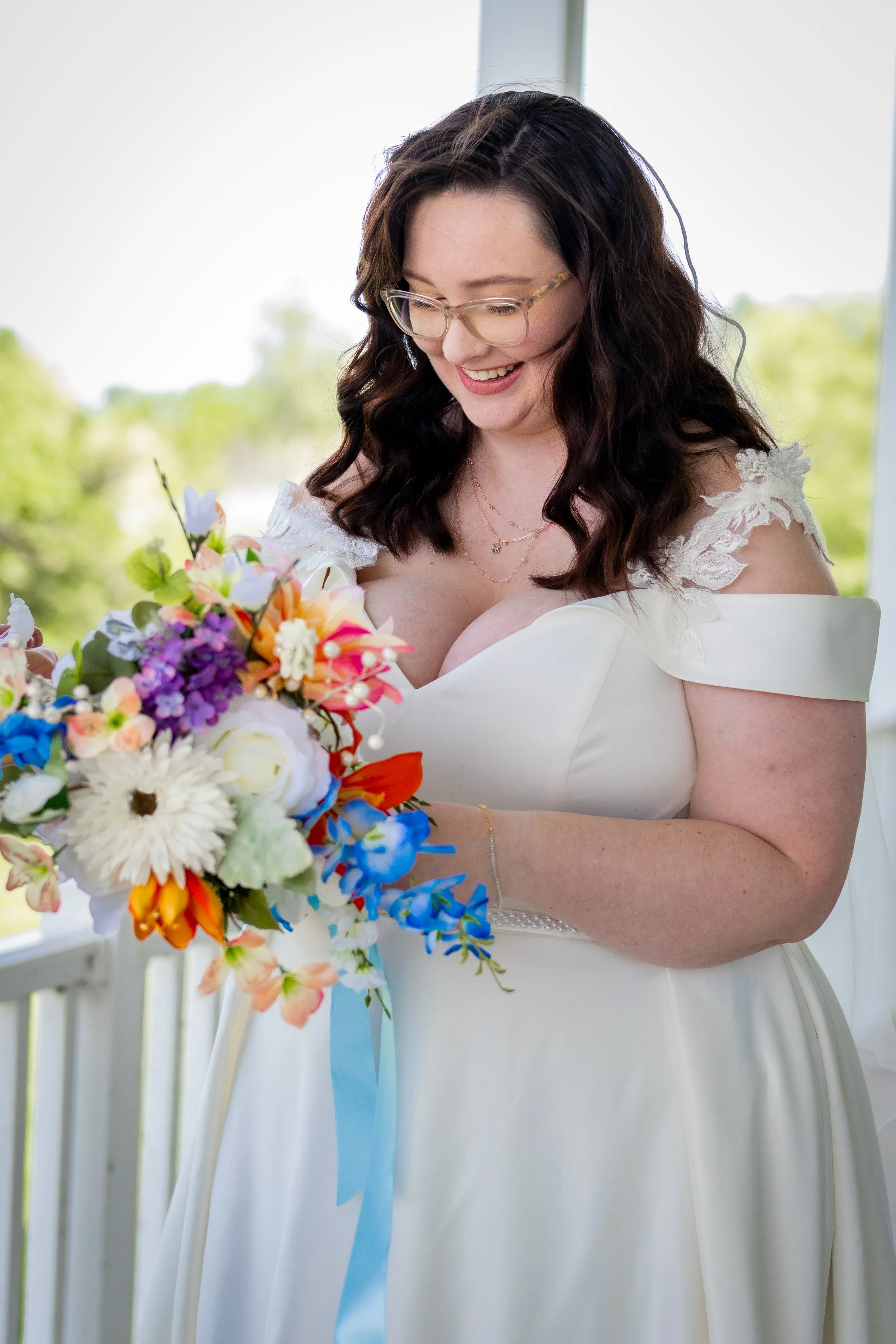 A woman in a white wedding dress holding a colorful bouquet of flowers, smiling, with a blurred outdoor background.