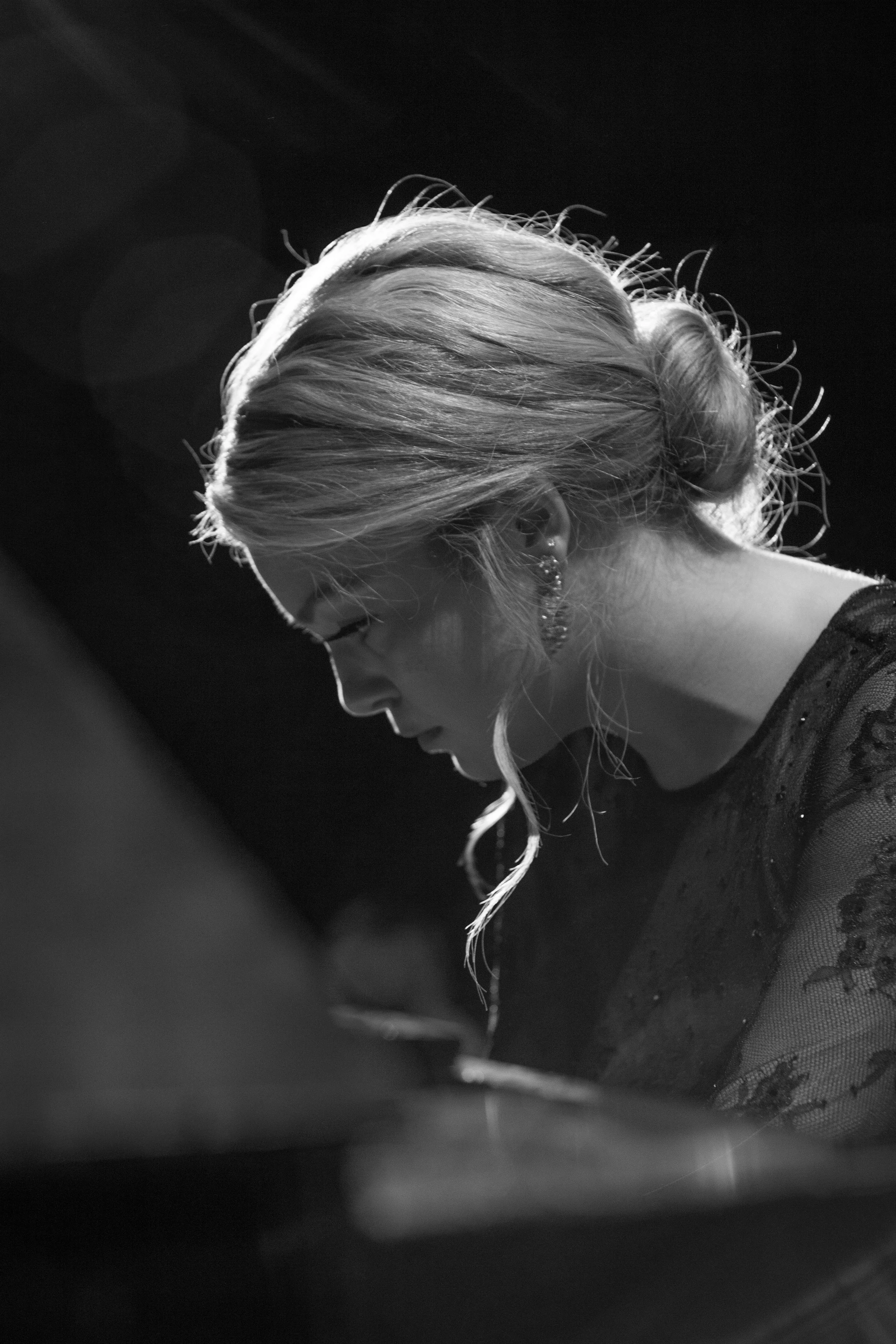 A black and white photo of a woman with styled hair and earrings, playing the piano with her head bowed down.