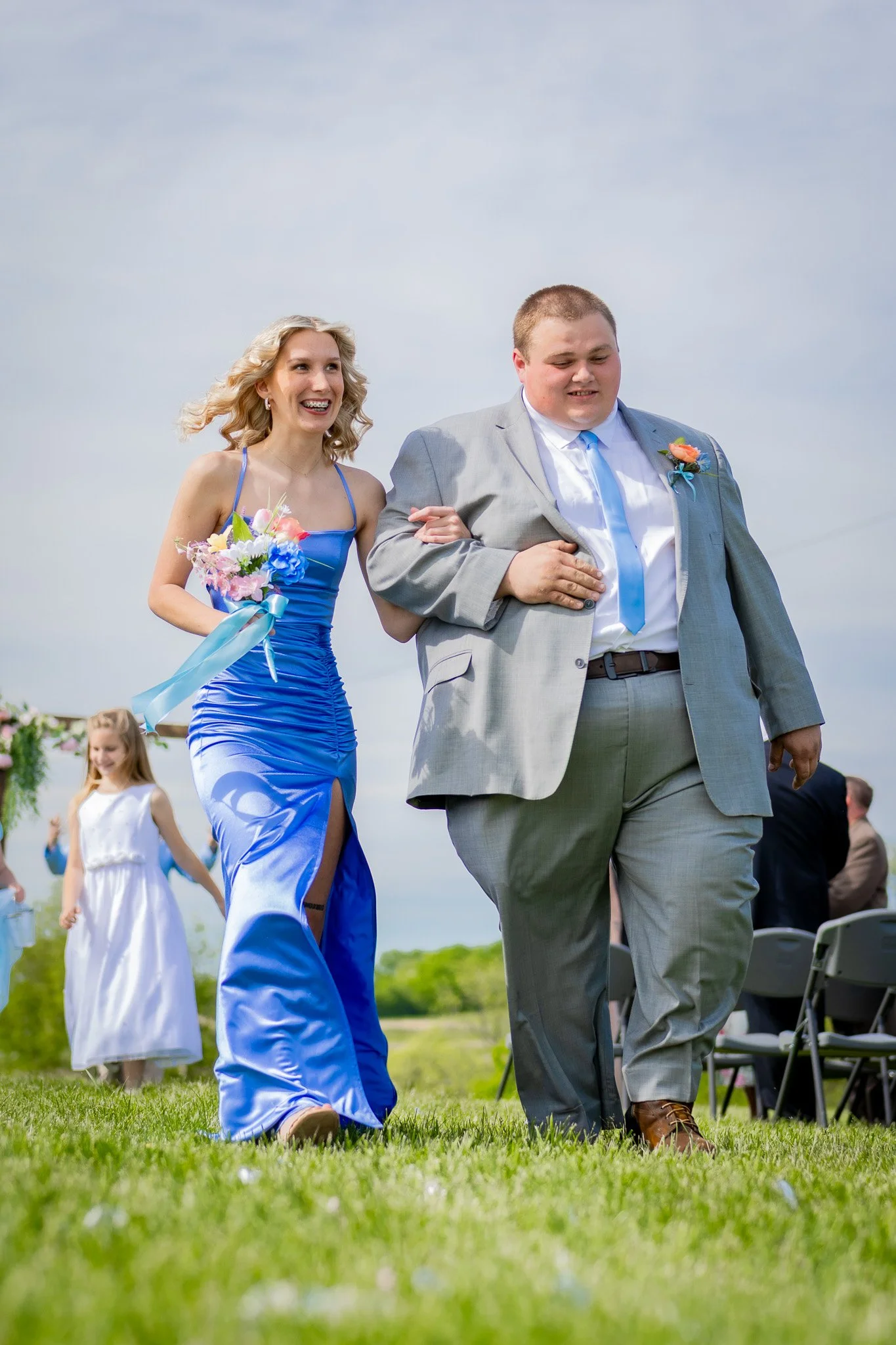 A woman in a blue dress holding a bouquet, walking with a man in a gray suit at an outdoor wedding ceremony, with guests seated in chairs and a girl in a white dress in the background.