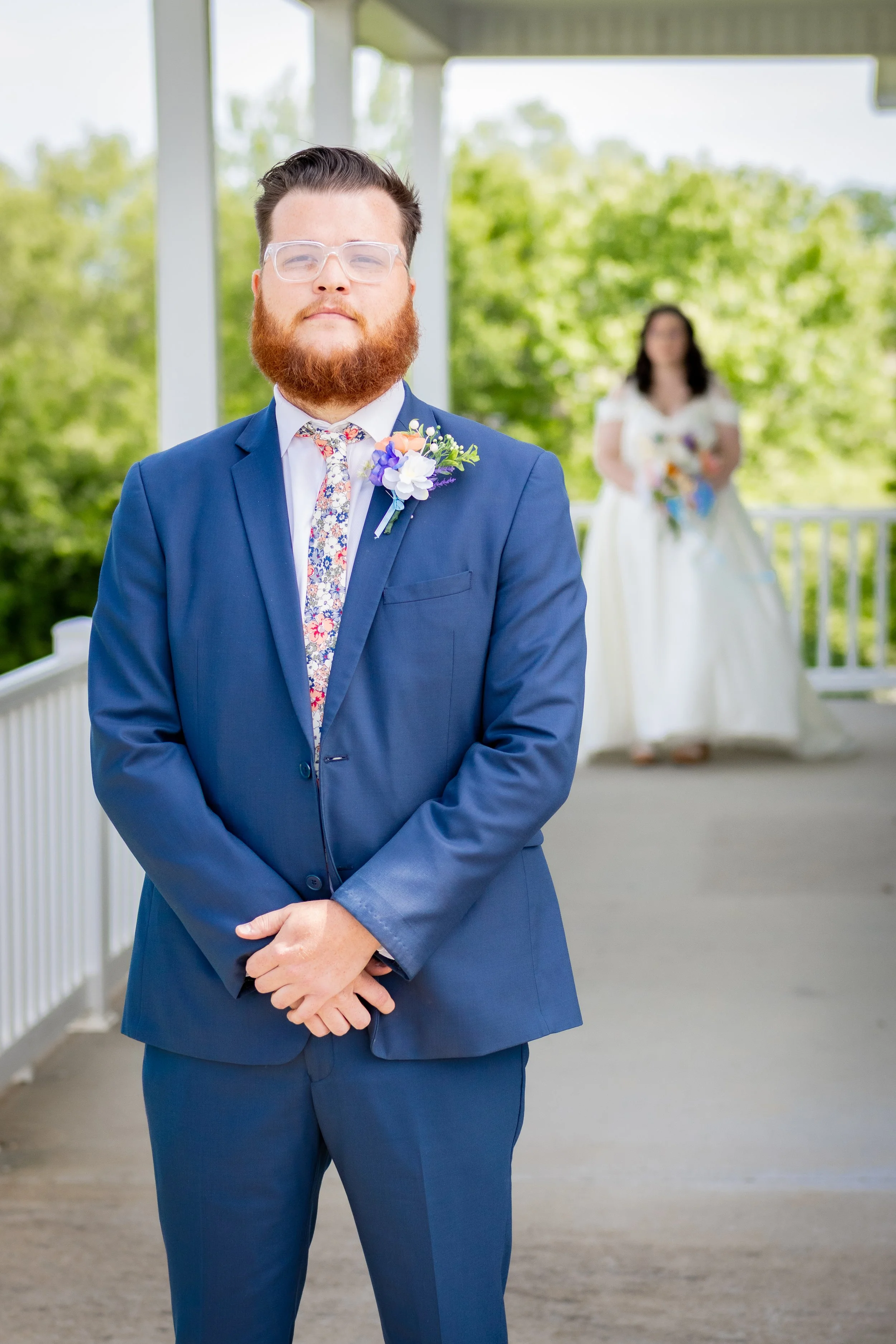 A man in a blue suit standing on a porch with a woman in a white wedding dress in the background holding a bouquet of flowers.