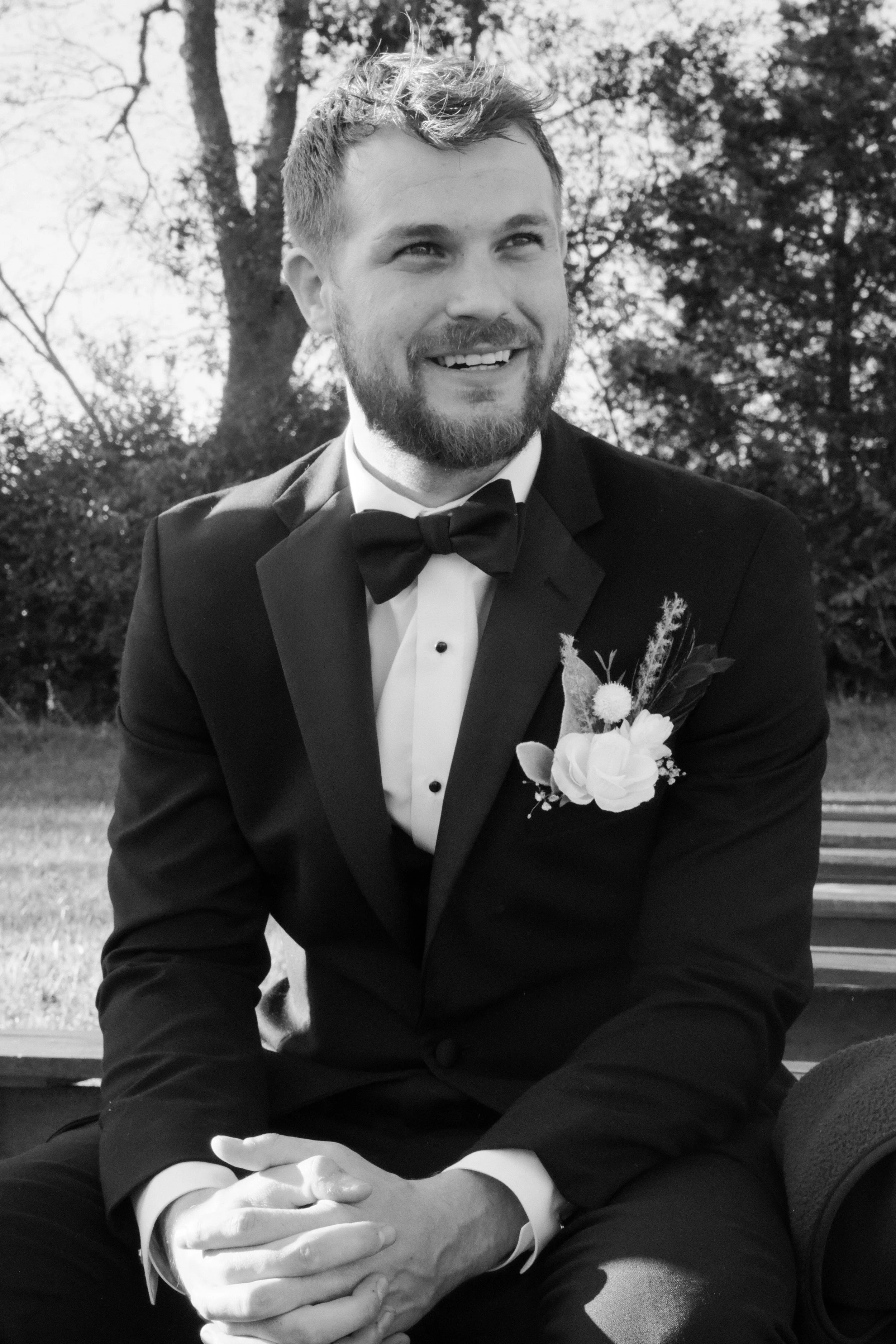 A man in a tuxedo with a bowtie and boutonniere, sitting outdoors on a park bench, smiling.