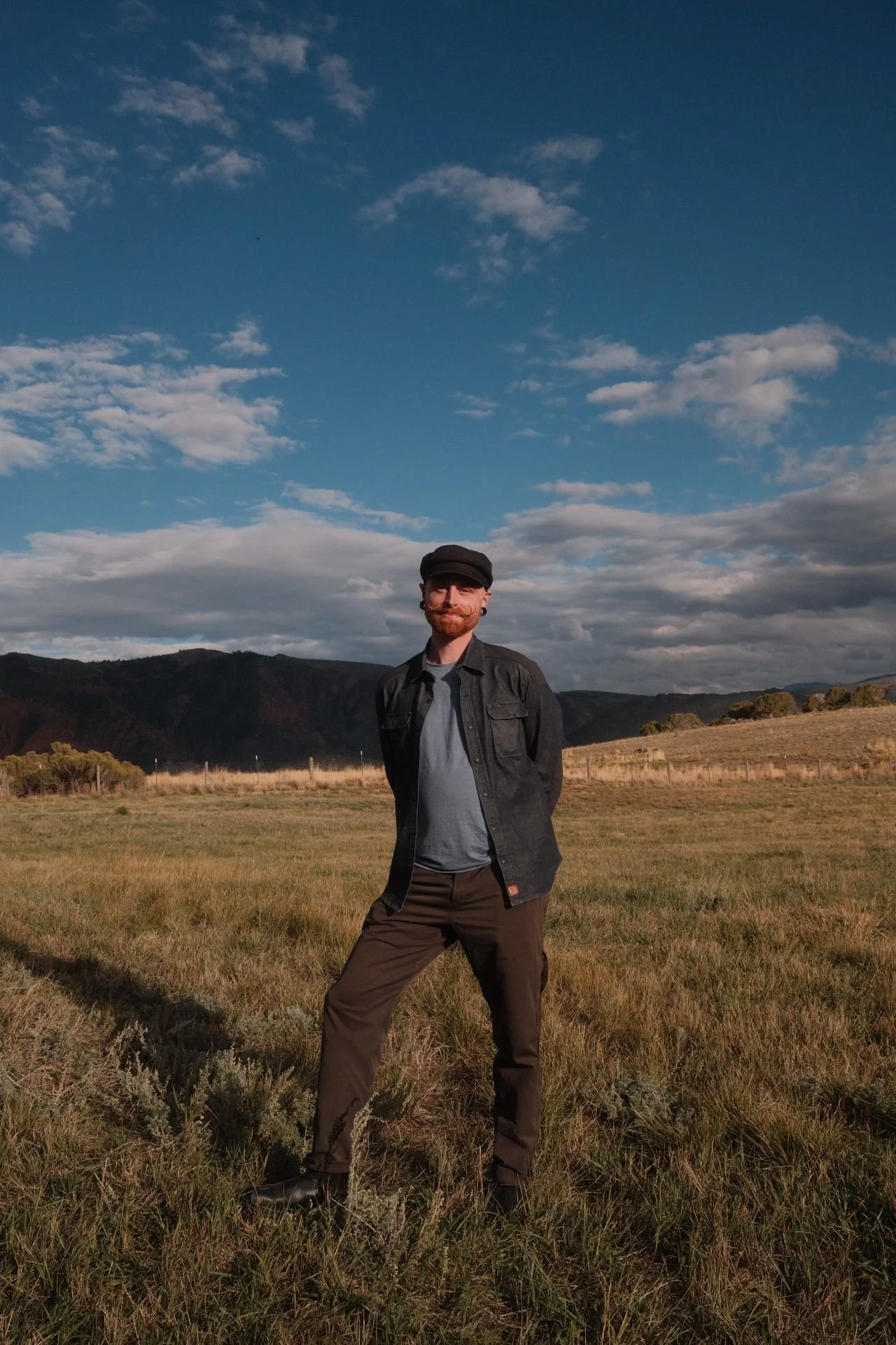 A man with a beard and mustache standing in a grassy field with his hands behind his back, wearing a black cap, black jacket, gray shirt, and brown pants, with a backdrop of hills and a partly cloudy sky.