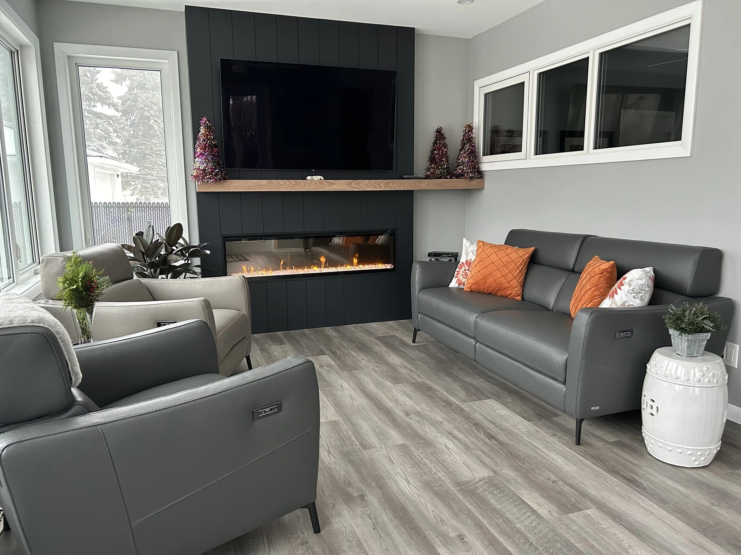 Modern living room with gray leather sofas, a white armchair, a fireplace with colorful decor, a mounted TV, and holiday decorations including small Christmas trees.